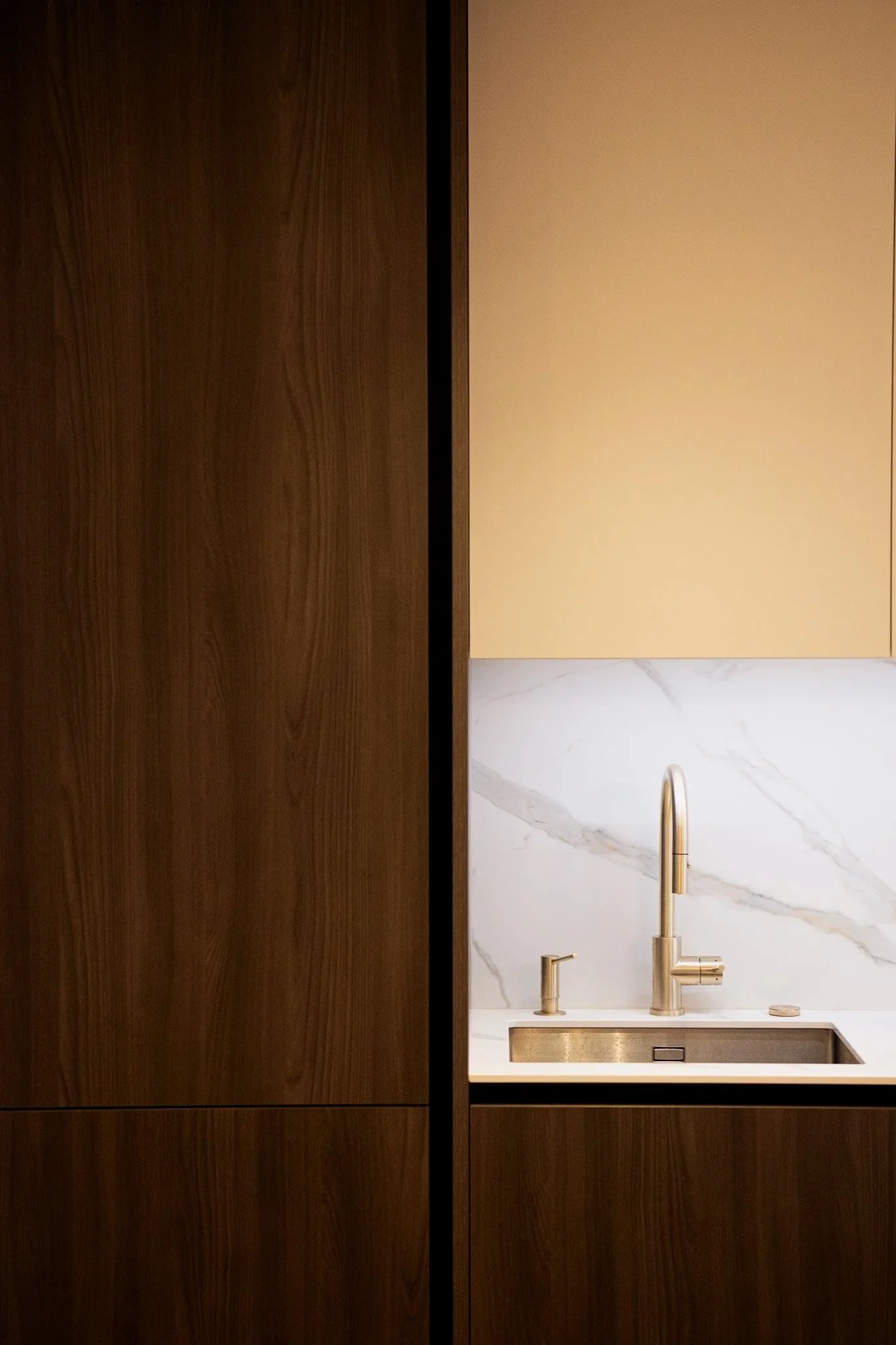 Une cuisine avec un meuble en bois foncé, un évier en métal doré, un robinet en métal doré, un fond de mur en marbre blanc avec veines grises et un placard de couleur beige.