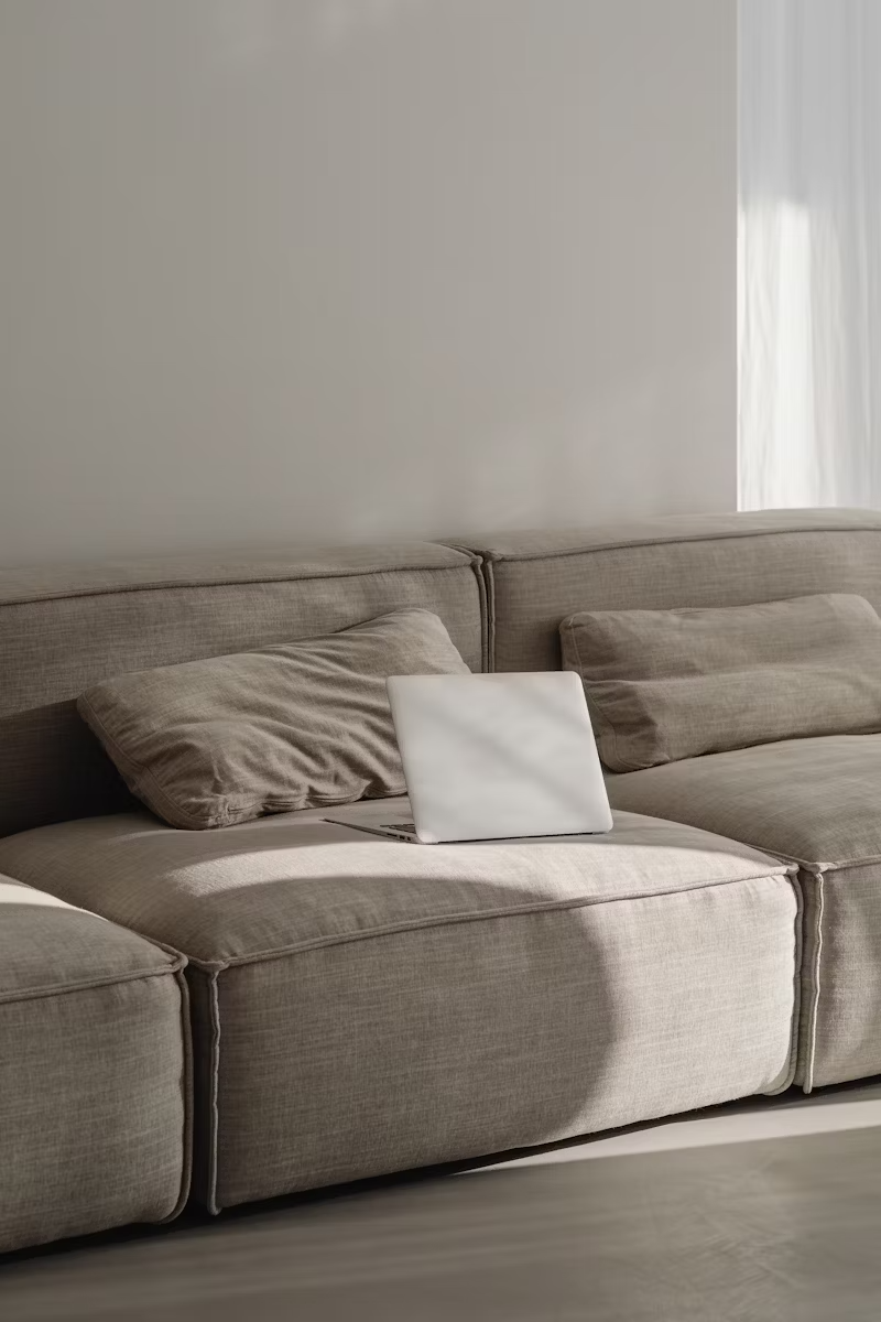 Canapé beige avec un coussin et un ordinateur portable fermé dessus, dans une pièce éclairée naturellement.