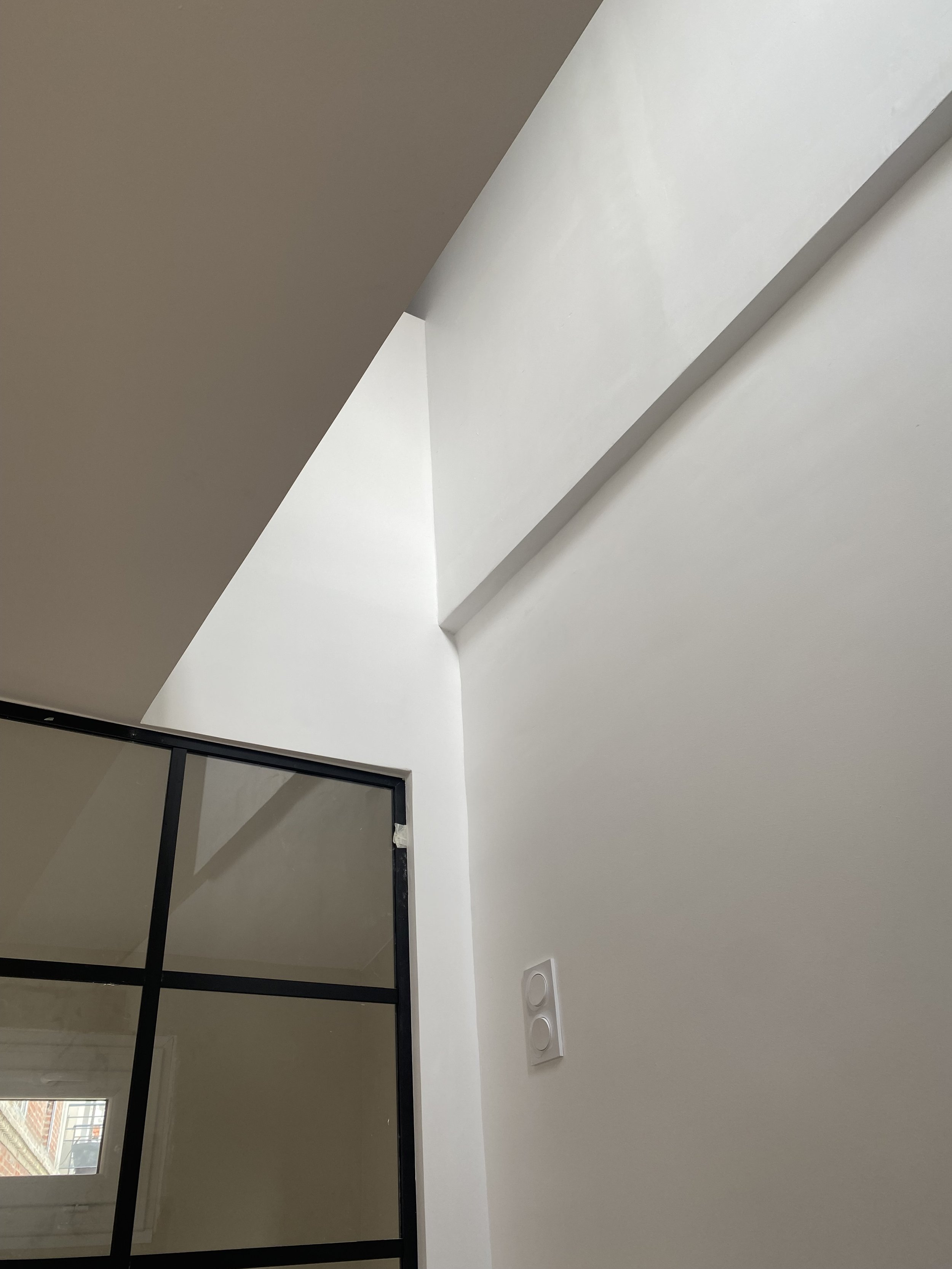 Vue intérieure d'un mur blanc avec une cloison en verre avec cadre noir, un interrupteur double blanc, et une zone de plafond avec une ouverture ou un espace vide, dans une pièce moderne.