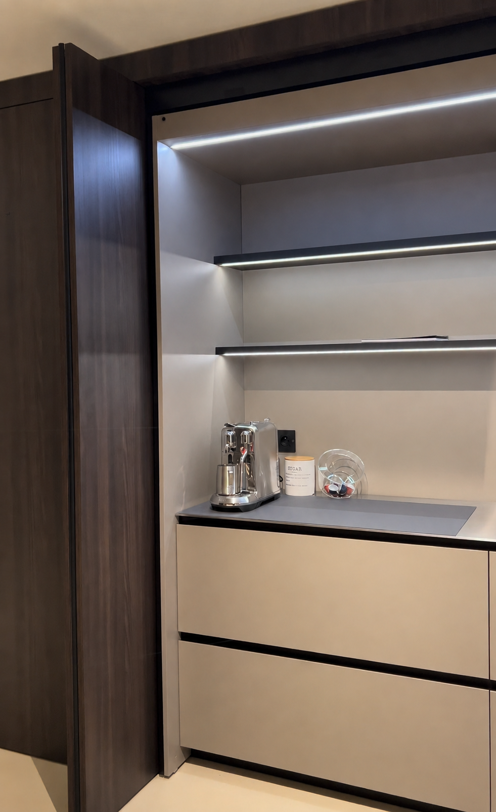 Une cuisine moderne avec un panneau en bois sombre, un plan de travail gris clair, une machine à café en acier inoxydable, un pot blanc avec inscription 'SUGAR', une boîte en verre contenant des capsules de café, et des étagères éclairées en arrière-