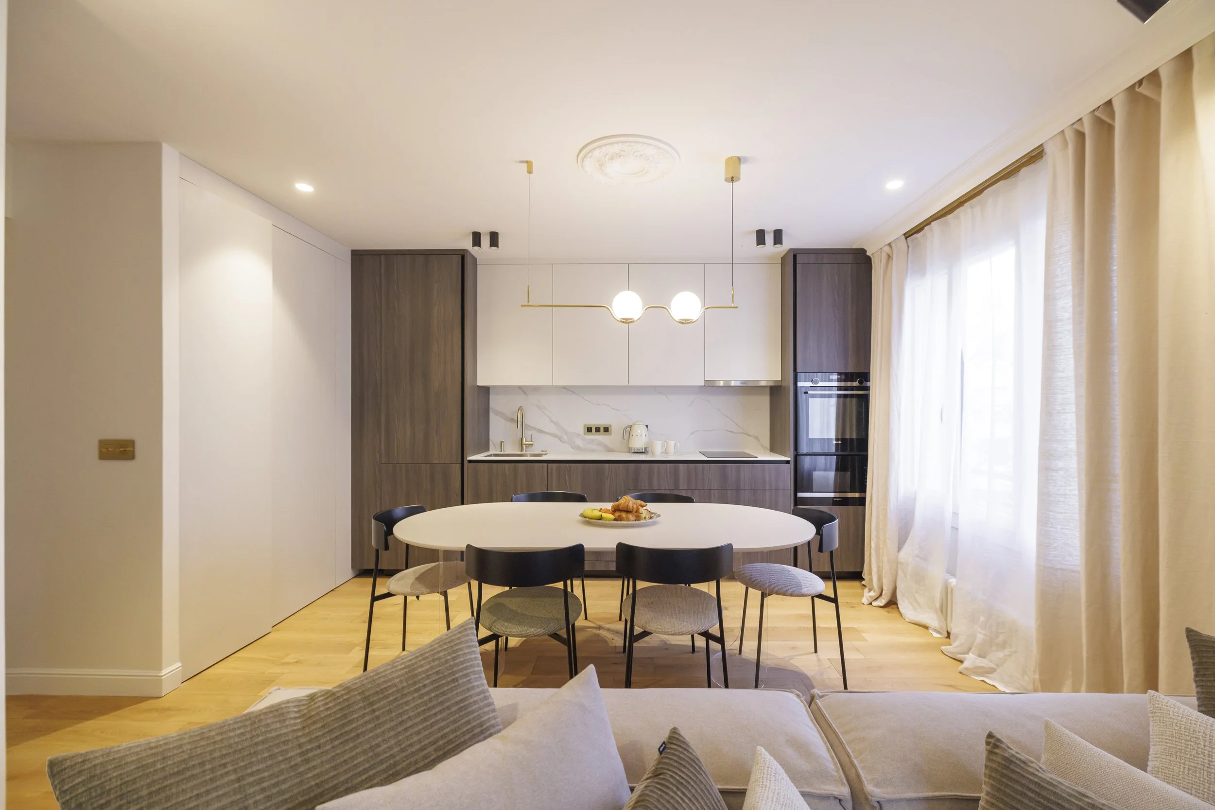 Intérieur d'une cuisine moderne avec une table rondes, des chaises noires, des armoires en bois foncé et blanche, un plan de travail blanc avec une cuisinière électrique, et un éclairage suspendu avec deux globes lumineux.