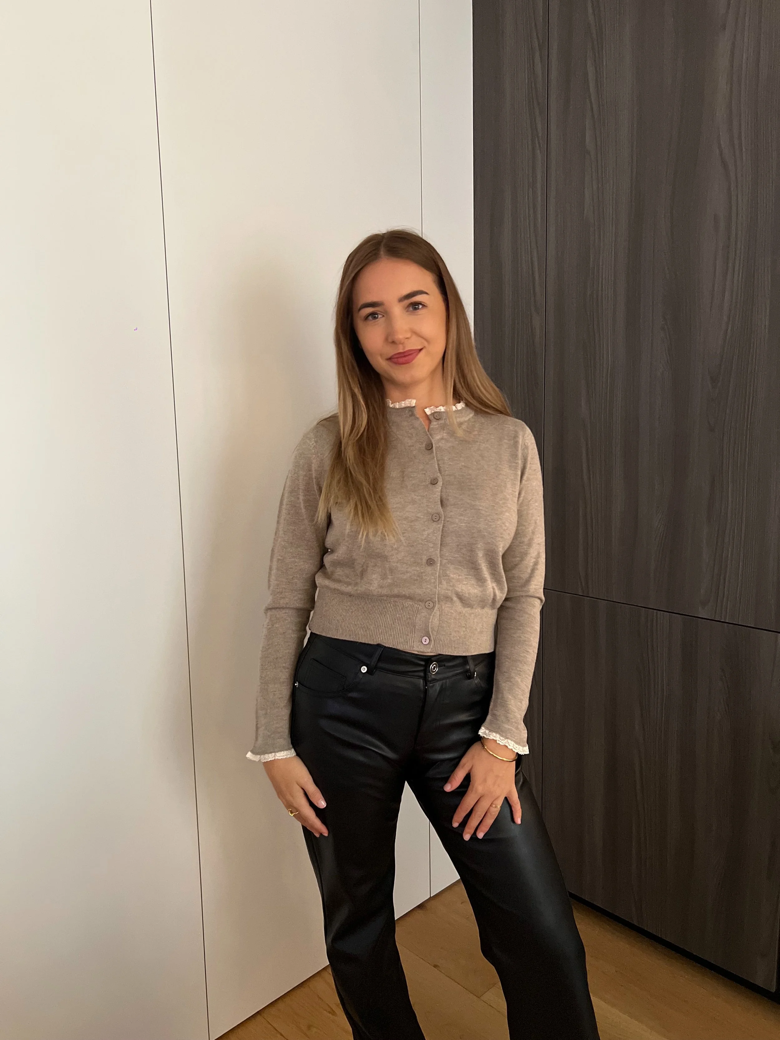 Une jeune femme dans une pièce moderne, portant un pull beige, un pantalon en cuir noir, et souriant, posant devant un mur blanc et un panneau en bois foncé.