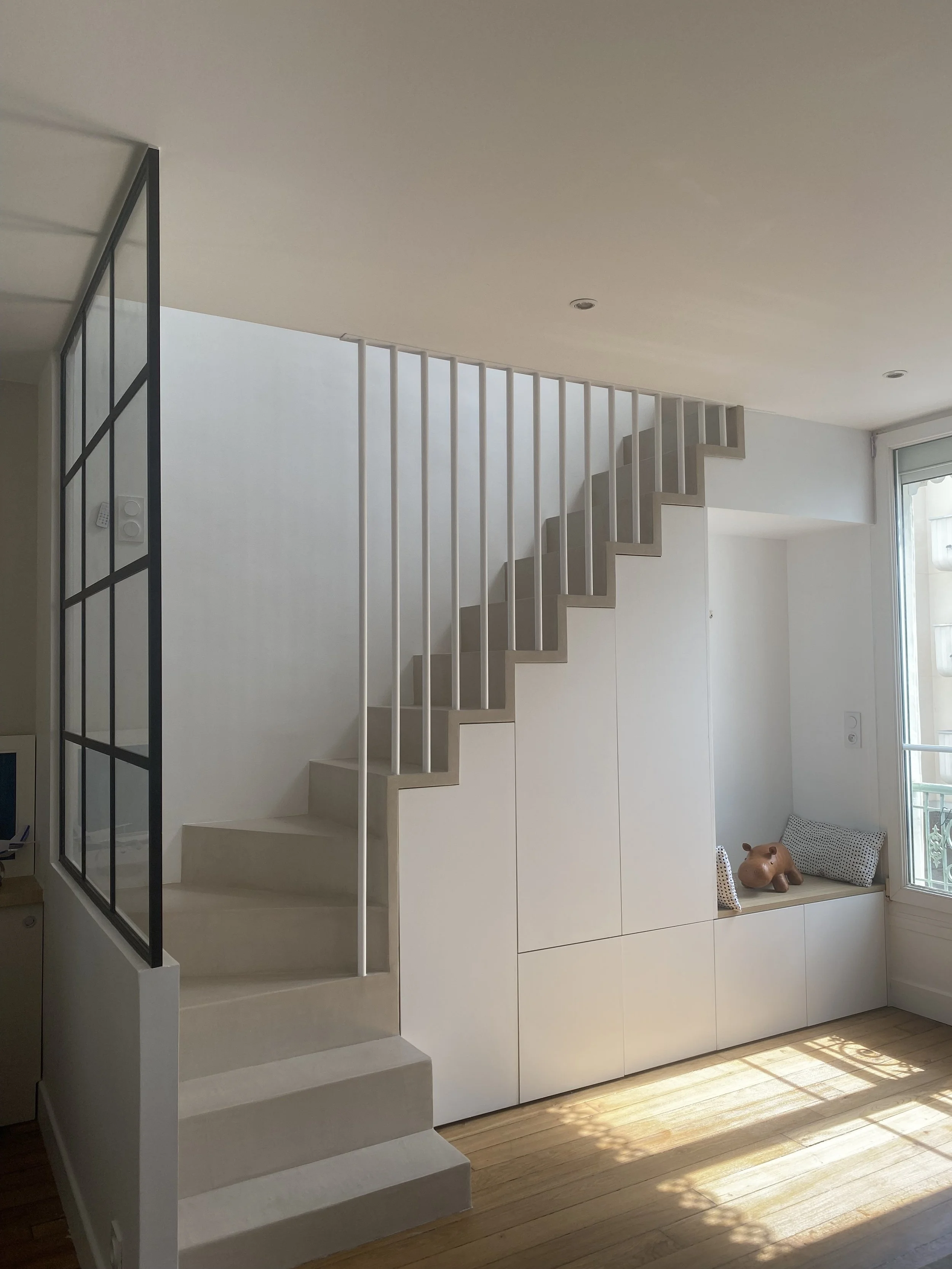 Intérieur moderne avec un escalier blanc en colimaçon, une grande fenêtre laissant entrer de la lumière naturelle, un mur blanc avec des rangements intégrés, et une étagère avec un coussin et une poupée en forme de chien en bas près de la fenêtre.