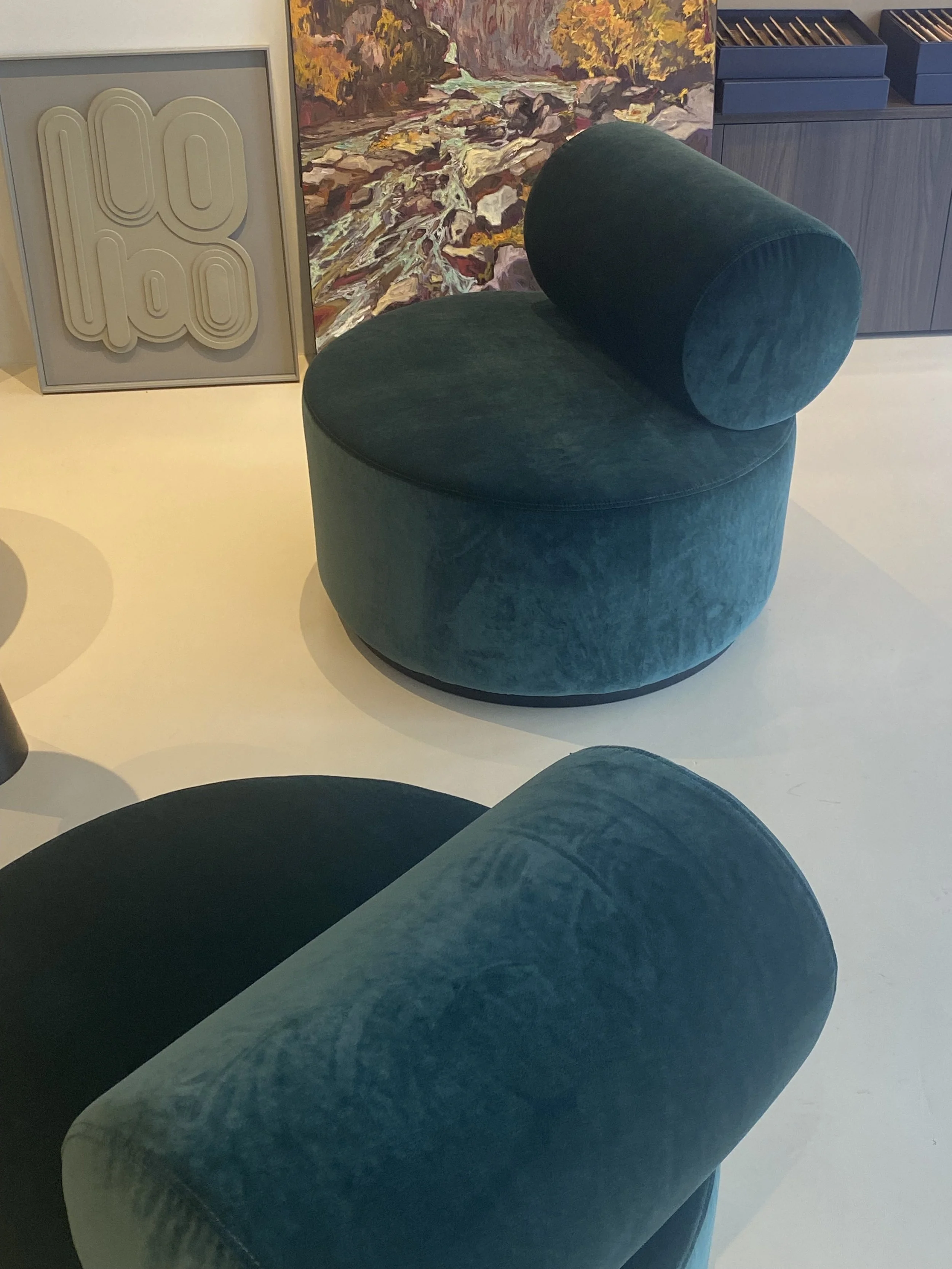 Deux fauteuils rembourrés en velours bleu, l'un en forme de cercle avec un coussin cylindrique, et l'autre en forme de demi-cercle, dans un espace décoratif avec une peinture artistique en arrière-plan.