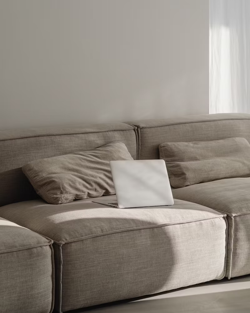 Un canapé beige avec des coussins et un ordinateur portable posé dessus dans un salon lumineux avec des rideaux blancs.