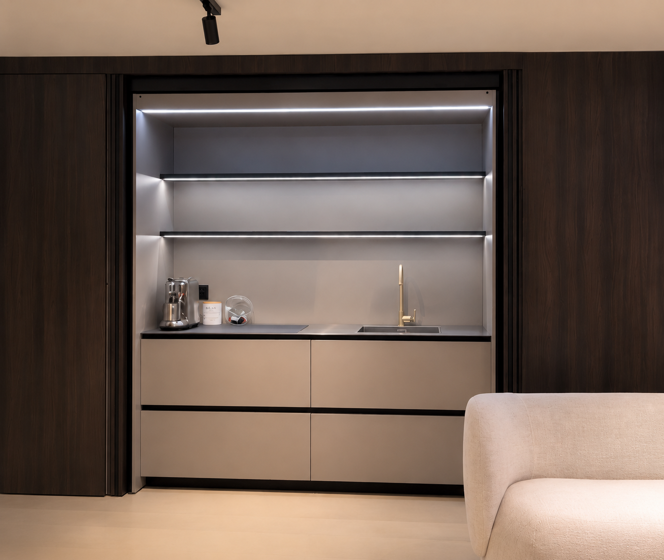 Une kitchenette moderne avec des armoires beige, un contour noir, un évier avec un robinet doré, et des étagères éclairées par des bandes lumineuses LED, dans un meuble encastré en bois foncé.