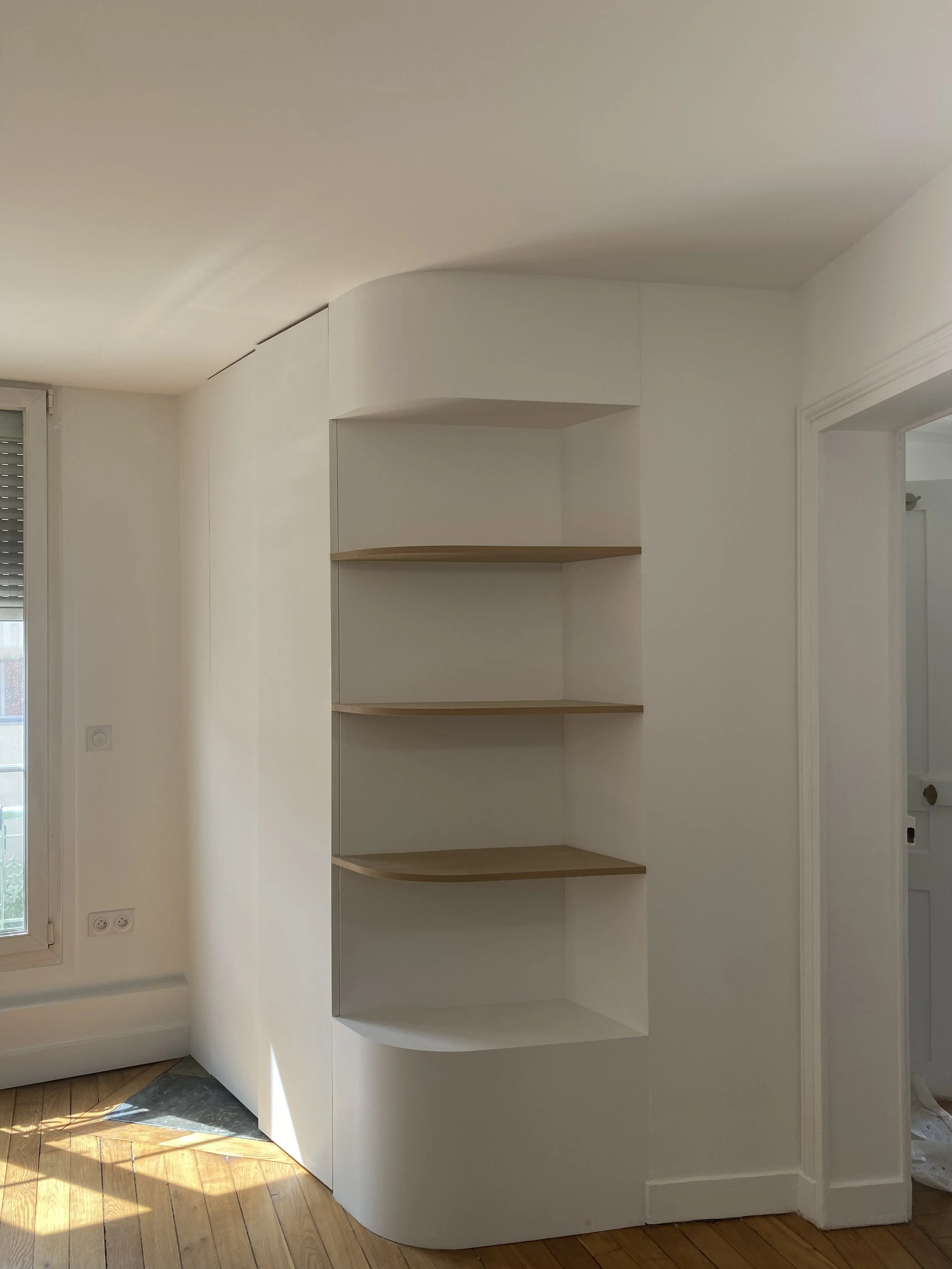 Un mur blanc avec une étagère courbée en bois clair dans une pièce lumineuse avec un sol en bois et une fenêtre.