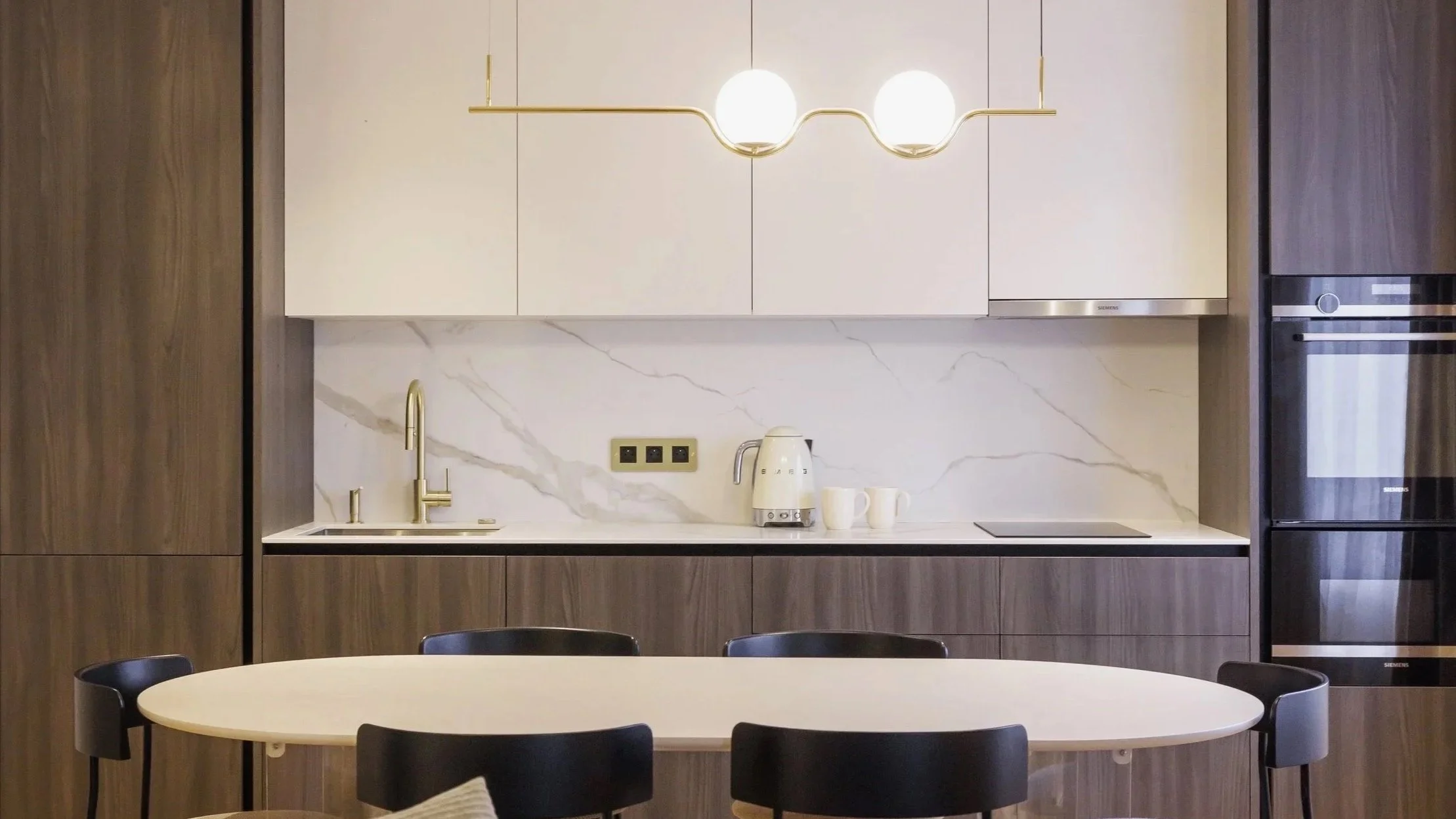 Cuisine moderne avec un plan de travail blanc, des armoires en bois sombre, un évier avec un robinet en or, une cafetière blanche, une plaque de cuisson noire, une tablette avec trois tasses blanches, un éclairage suspendu avec deux globes lumineux