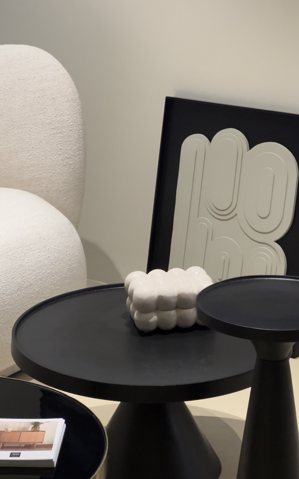 Décoration intérieure avec un tableau noir avec une œuvre abstraite blanche, une sculpture en céramique blanche en forme de bulles, un fauteuil blanc en tissu texturé et deux tables noires de styles différents.