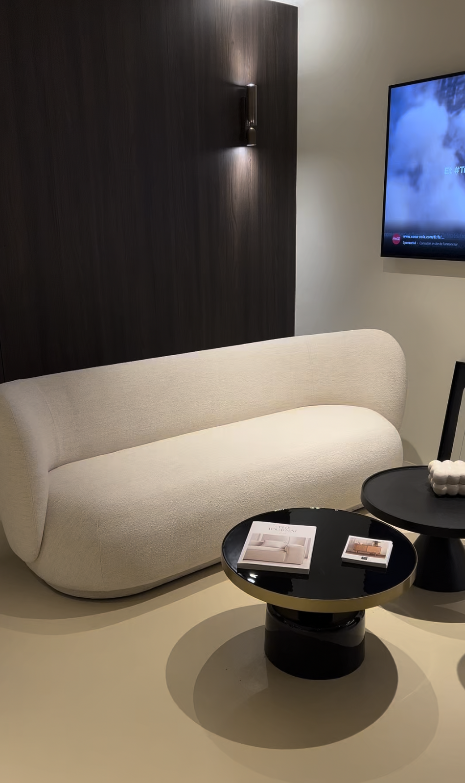 Salon moderne avec canapé beige, deux tables basses noires avec magazines, mur en bois foncé avec appliqué lumineux, télévision murale affichant une image de paysage.