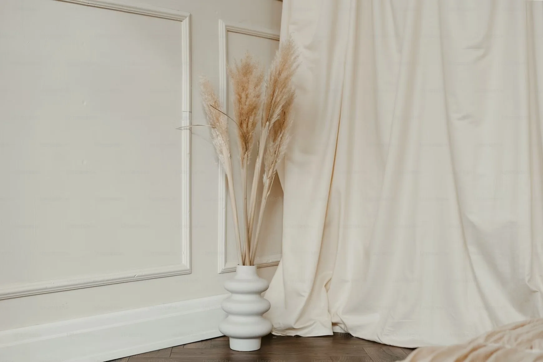 Vase blanc avec des pampas feuillus beiges, placée à côté d'un mur crème avec moulures et d'un rideau crème