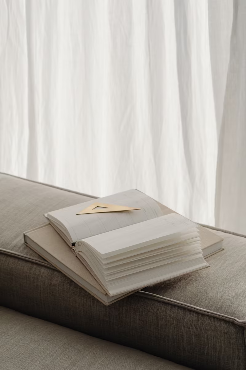 Un cahier ouvert avec des pages blanches repose sur un canapé, avec une règle en plastique posée dessus, devant un rideau blanc léger.