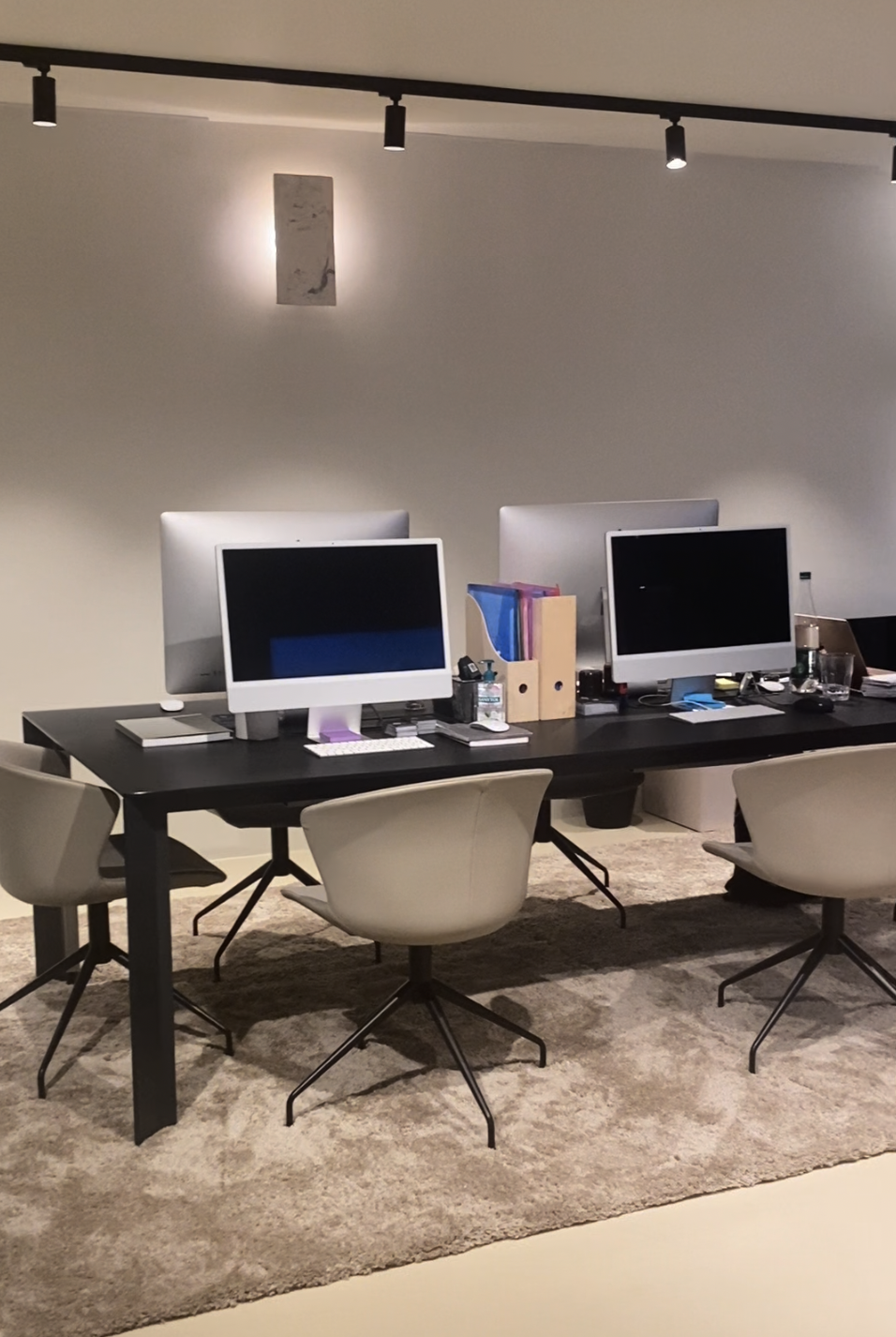 Bureau moderne avec trois ordinateurs ergonomiques, des chaises grises, des dossiers colorés, des objets de bureau, et une décoration minimaliste, éclairé par un système d'éclairage au plafond.