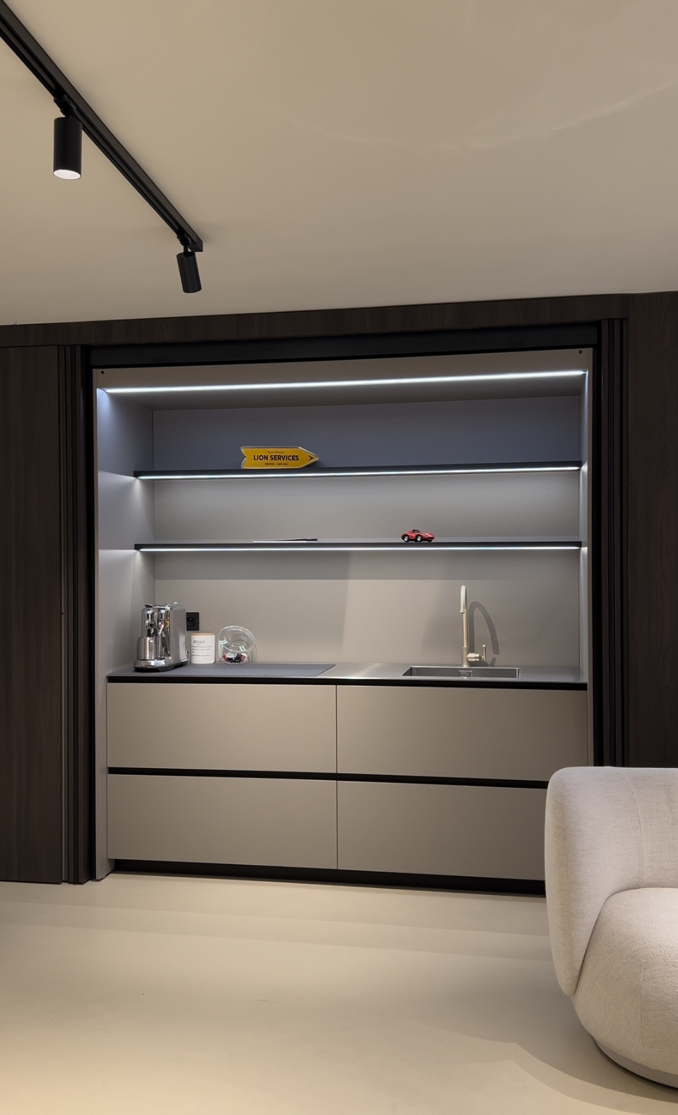 Une petite kitchenette moderne avec un comptoir, un évier, une cafetière, un pot contenant des objets, une étagère avec une petite voiture rouge, une lampe de plafond, et un fauteuil beige partiellement visible à droite.