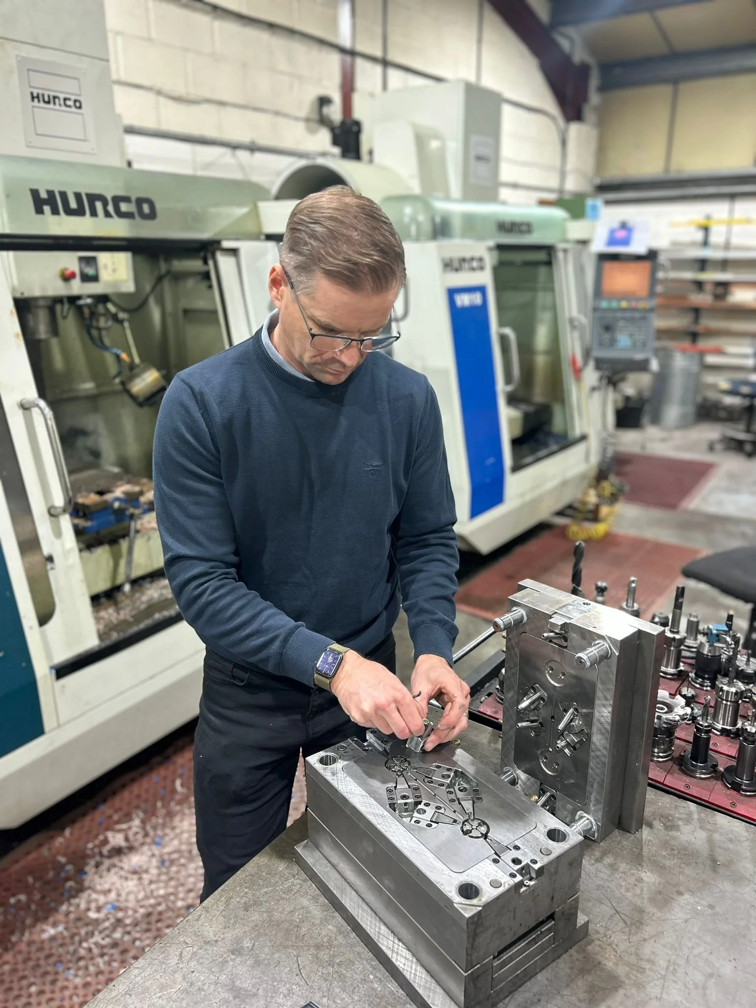 A man working on a metal mold or fixture in an industrial workshop, with CNC machines in the background.