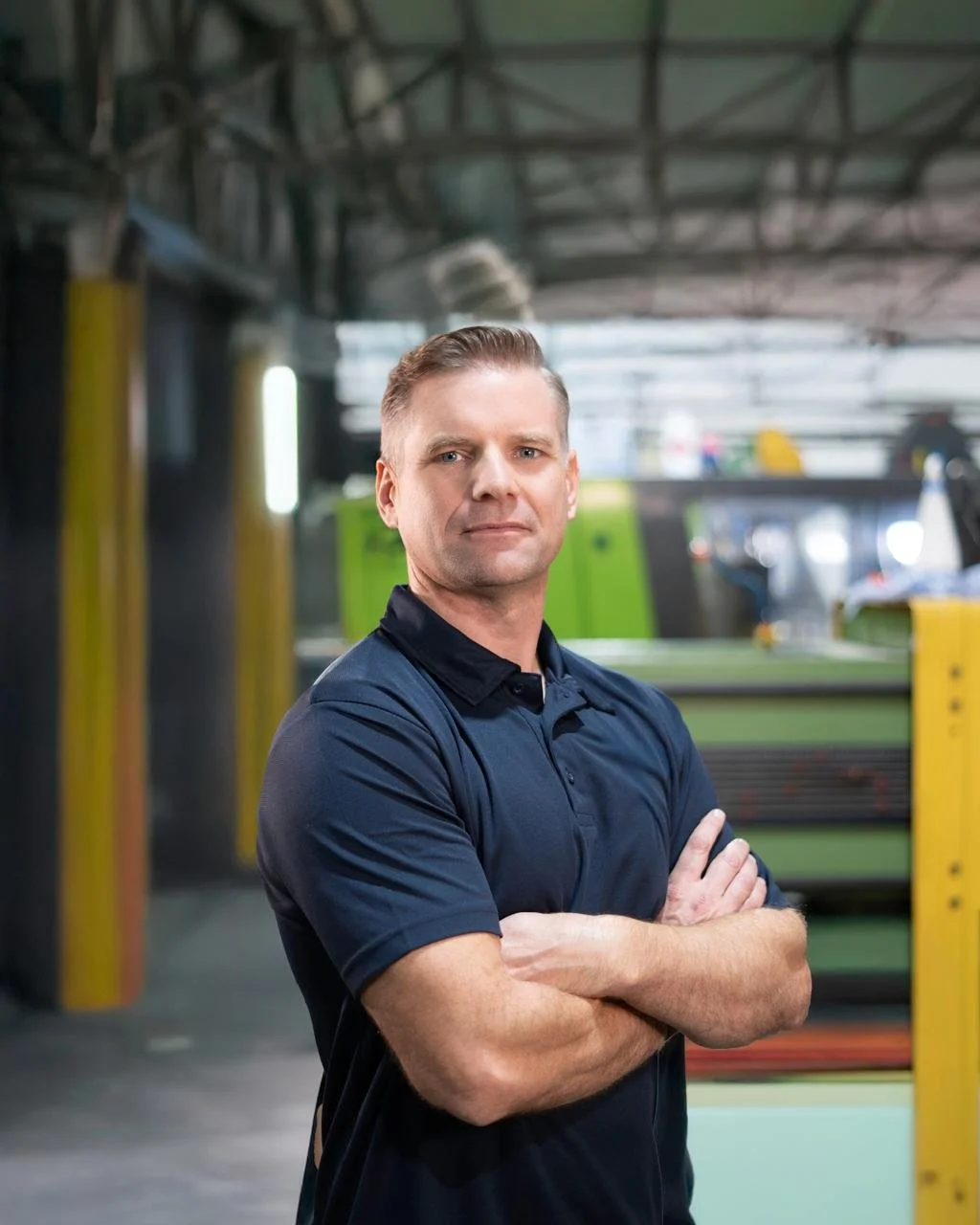 A confident man with crossed arms standing in an industrial or manufacturing setting with machinery in the background.