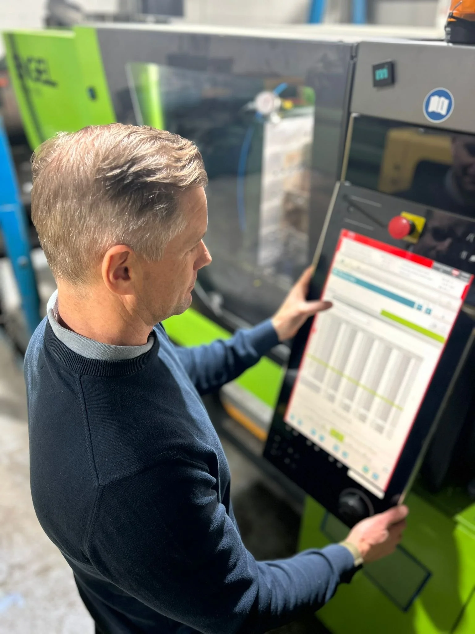 A man with gray hair wearing a blue sweater using a touchscreen control panel on industrial equipment in a factory or workshop.