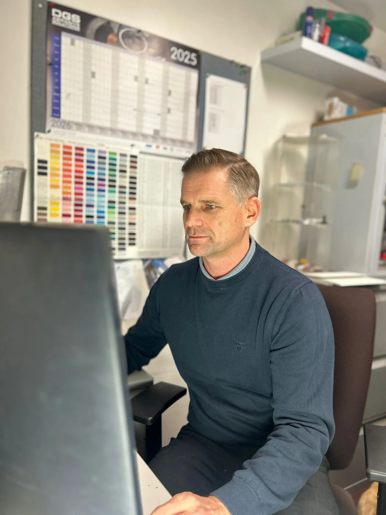A man sitting at a desk working on a computer in an office with posters, calendars, and shelves in the background.