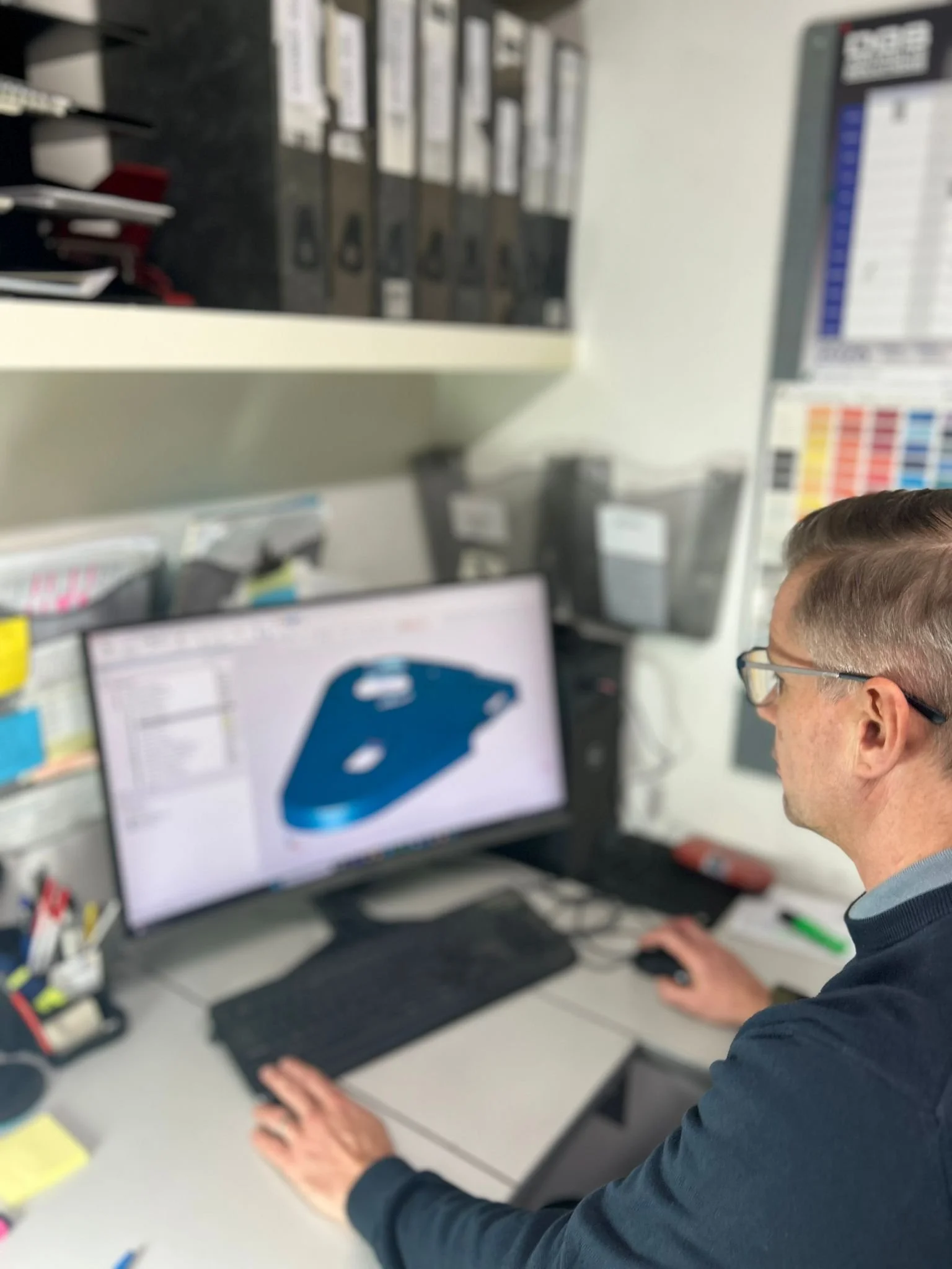 A man working at a desk on a computer, viewing a 3D model of a mechanical part on the screen, surrounded by office supplies and shelves.