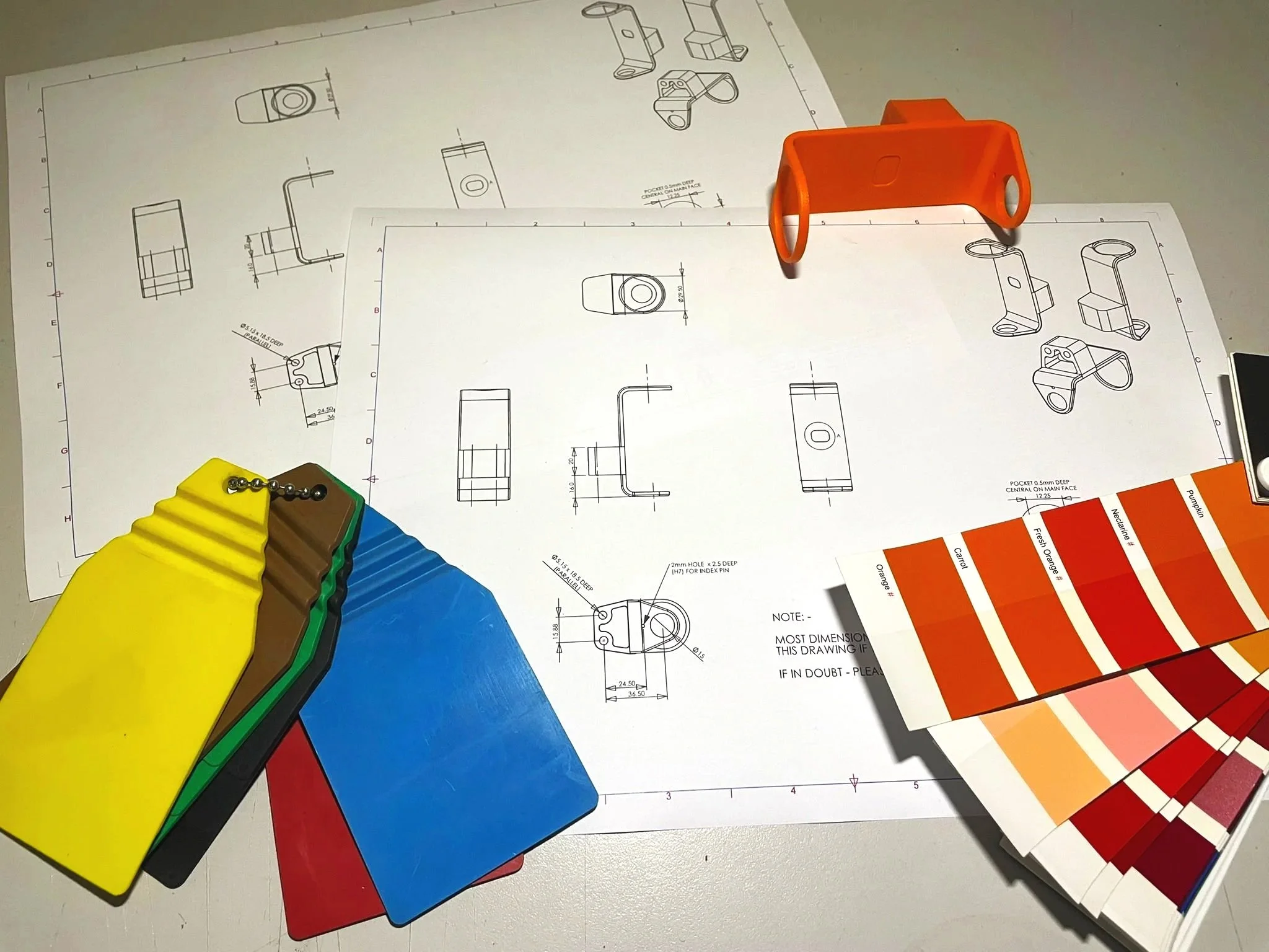 Technical drawing and color samples for a mechanical or design project, including color swatches, engineering blueprints, and small metal keychain-like objects.