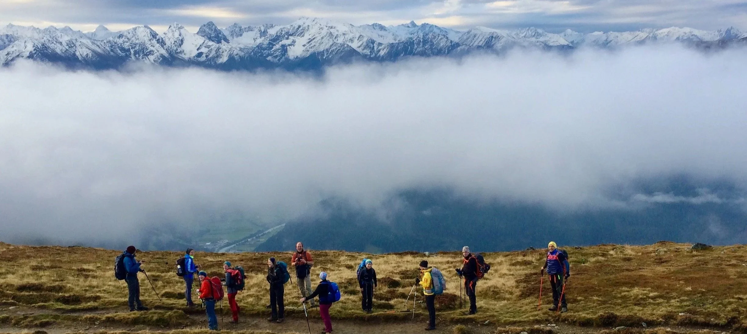 Gruppe von Wanderern auf einer Bergwiese mit Blick auf schneebedeckte Berge im Hintergrund, umgeben von Wolken.