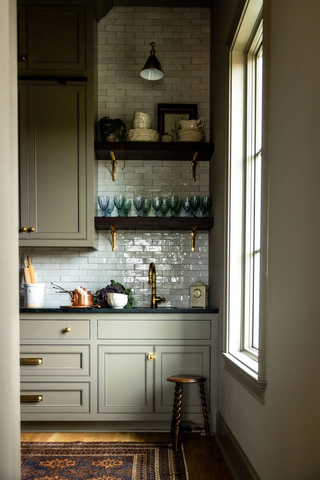 A cozy kitchen corner with sage green cabinets, white subway tile backsplash, and dark wooden open shelves holding dishes, glasses, and framed pictures. A small wooden stool is beside the cabinets near a window, which lets in natural light. Wolff Dur