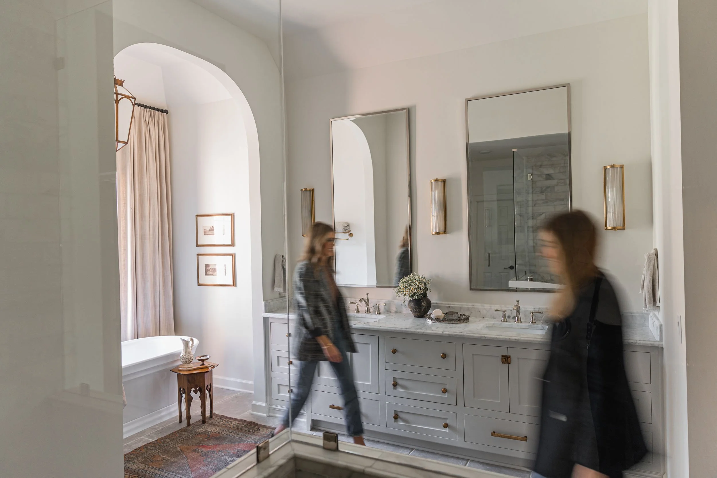 A modern bathroom with large mirrors, double sinks, and a women walking past. Wolff Duran Studio Interior Design.