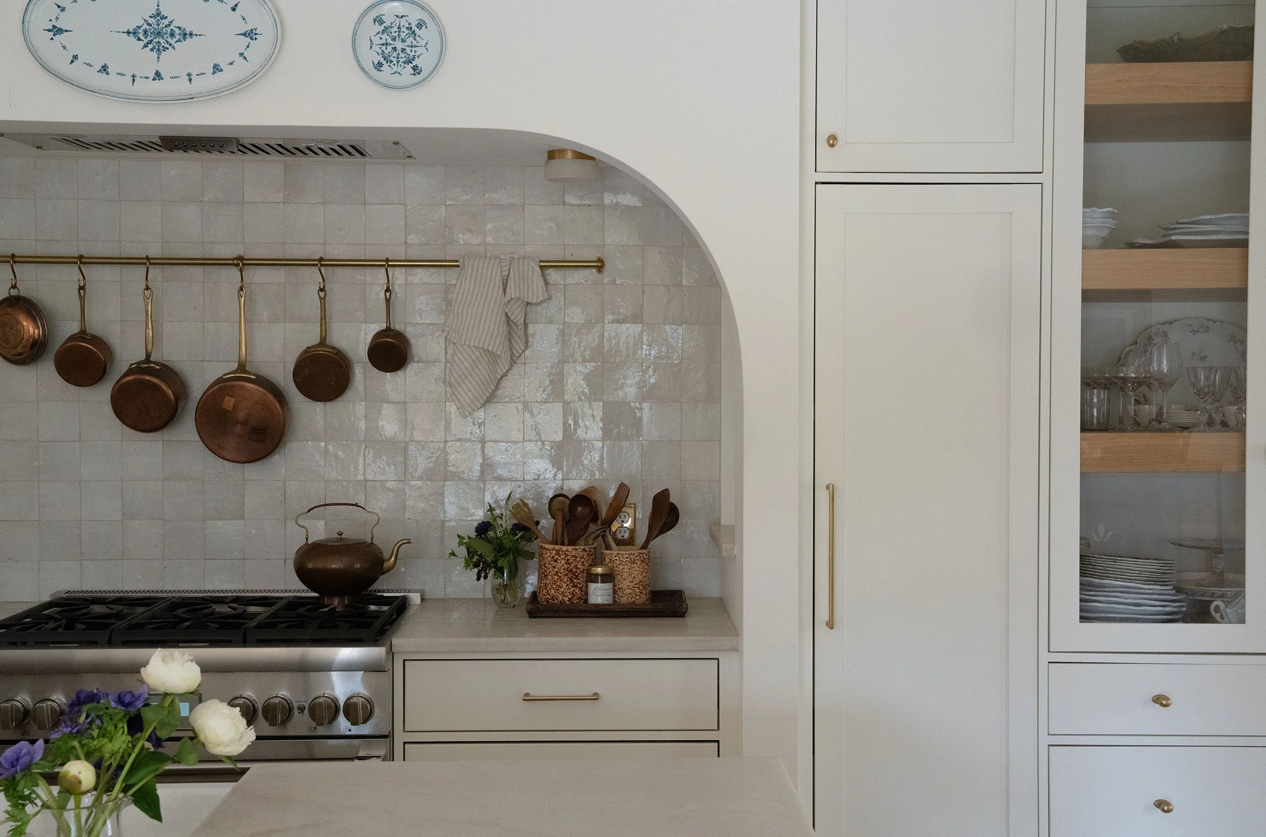 Kitchen with beige cabinets, a stove with a brown tea kettle, pots hanging on a rail, a vase with flowers, and open shelving with plates and glasses. Wolff Duran Studio Interior Design.