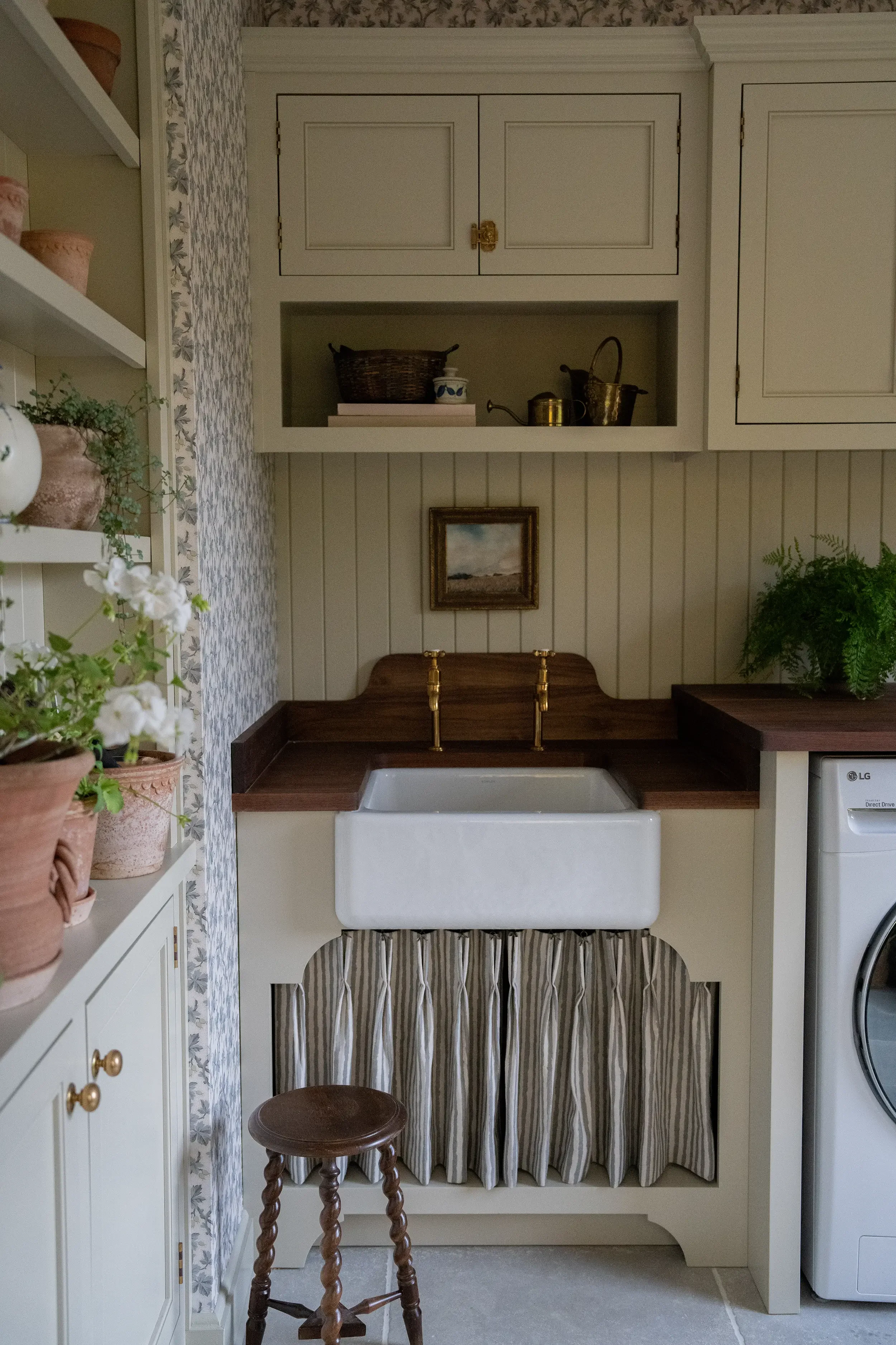 A vintage laundry room with a large farmhouse sink, brown wood countertop, white beadboard paneling, and open shelves with antique decor, plants, and baskets. Wolff Duran Studio Interior Design.