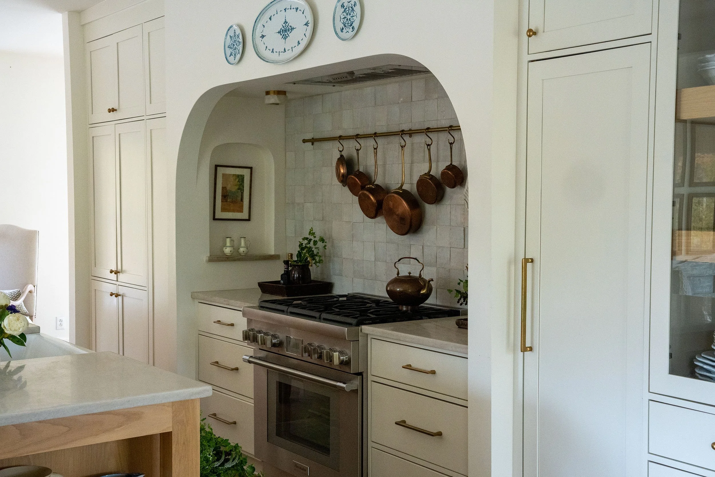 Kitchen with cream-colored cabinets, a stove, and a hanging copper pot rack, decorated with a wall clock and artwork. Wolff Duran Studio Interior Design.