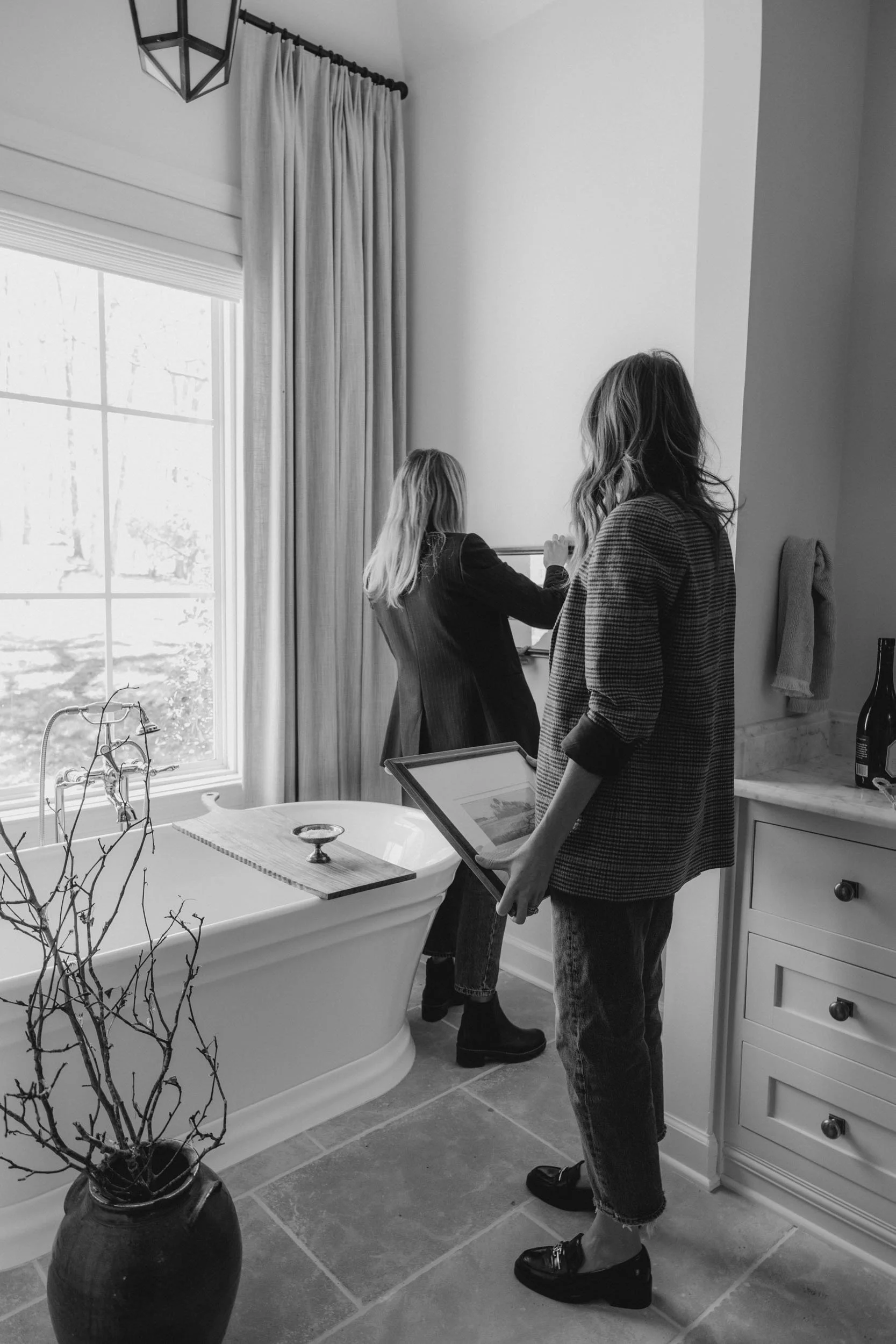 Two women standing in a bright bathroom, one adjusting a picture on the wall, the other holding a framed photo, with a large window, bathtub, and a vase with branches nearby.