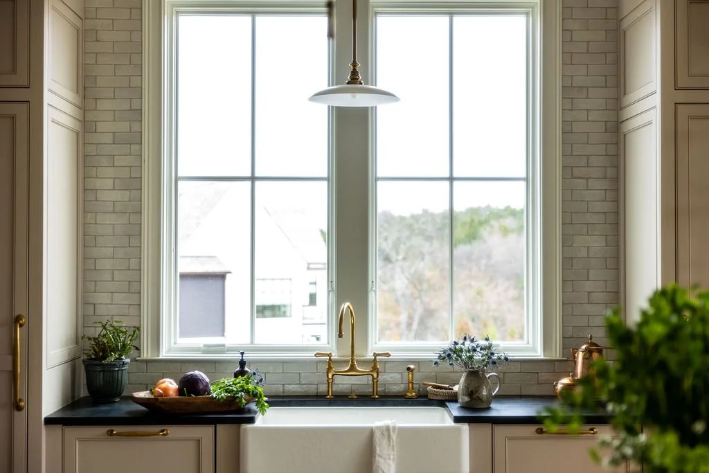 Kitchen sink area with large window, white brick backsplash, and cream cabinets with gold handles. There are plants, vegetables, and a flower arrangement on the black countertop. Wolff Duran Studio Interior Design.