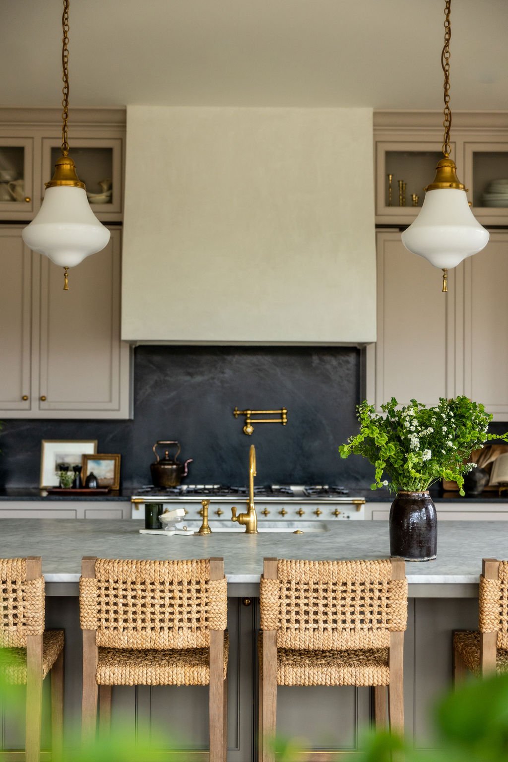 A modern kitchen with beige cabinetry, black marble backsplash, gold fixtures, two white pendant lights, a marble countertop island, and a black pot with green plants on it. Wolff Duran Studio Interior Design.