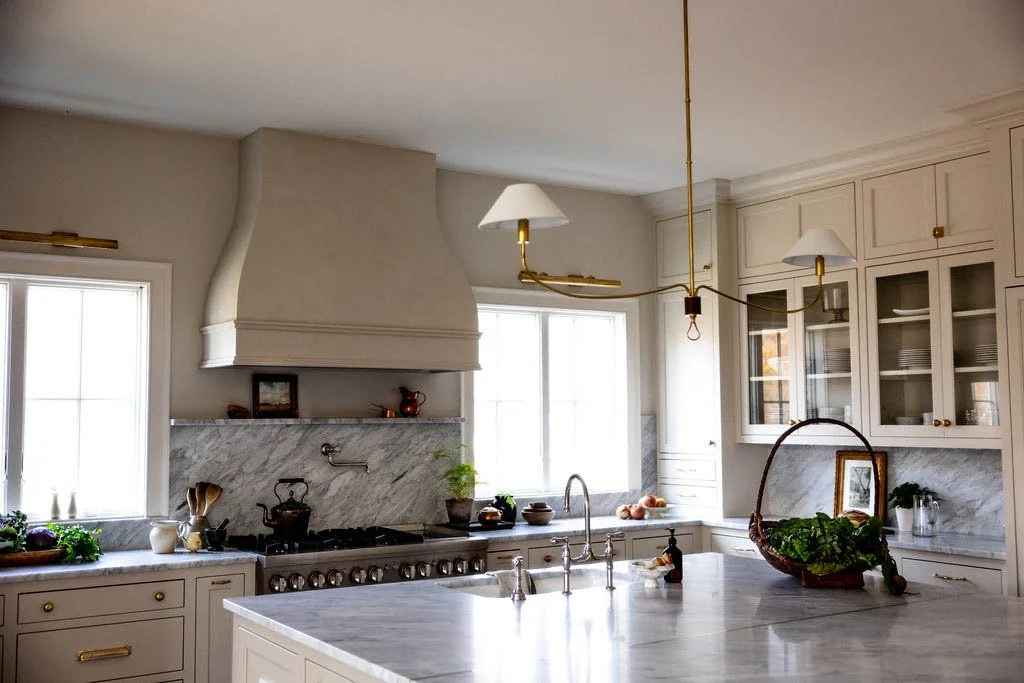 Bright kitchen with white cabinetry, marble countertops, and a large island. There is a vintage-style range hood above the stove, and a chandelier hanging over the island. Windows let in natural light, and there are plants and kitchenware on the countertops.