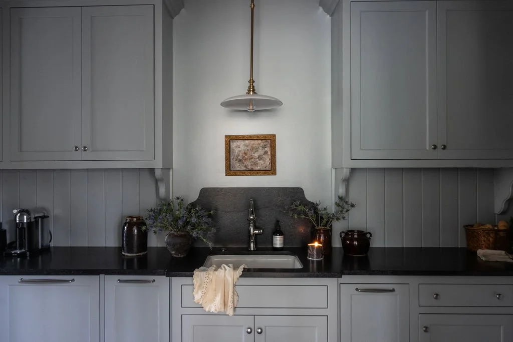 A kitchen with gray cabinets, a black countertop, and a small single sink. Decor includes two vases with greenery, a candle, and a small bottle. A hanging light fixture is above the sink, and a framed picture is on the wall behind it. There is a coff
