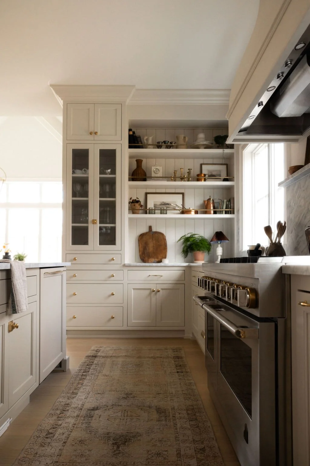 White kitchen with open shelves displaying dishes and decorative items, stainless steel oven, and a window letting in natural light. Wolff Duran Studio Interior Design.
