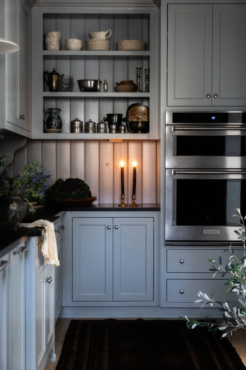 A cozy kitchen corner with open gray shelves holding ceramic bowls, cups, and vintage containers, illuminated by candlelight, with light gray cabinets, a built-in double oven, and a dark countertop decorated with plants and flowers.
