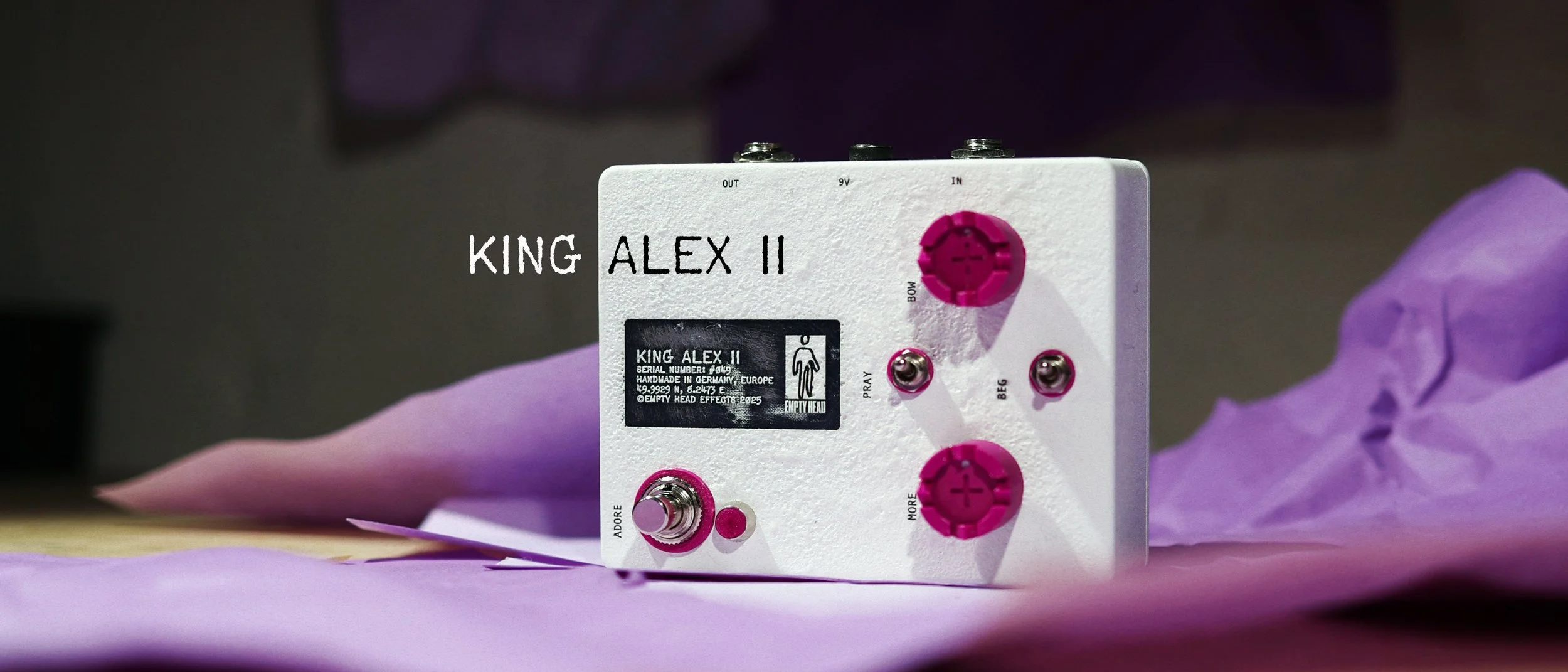A white guitar effects pedal with pink knobs surrounded by gold curly ribbons, featuring the text "KING ALEX II" in white.