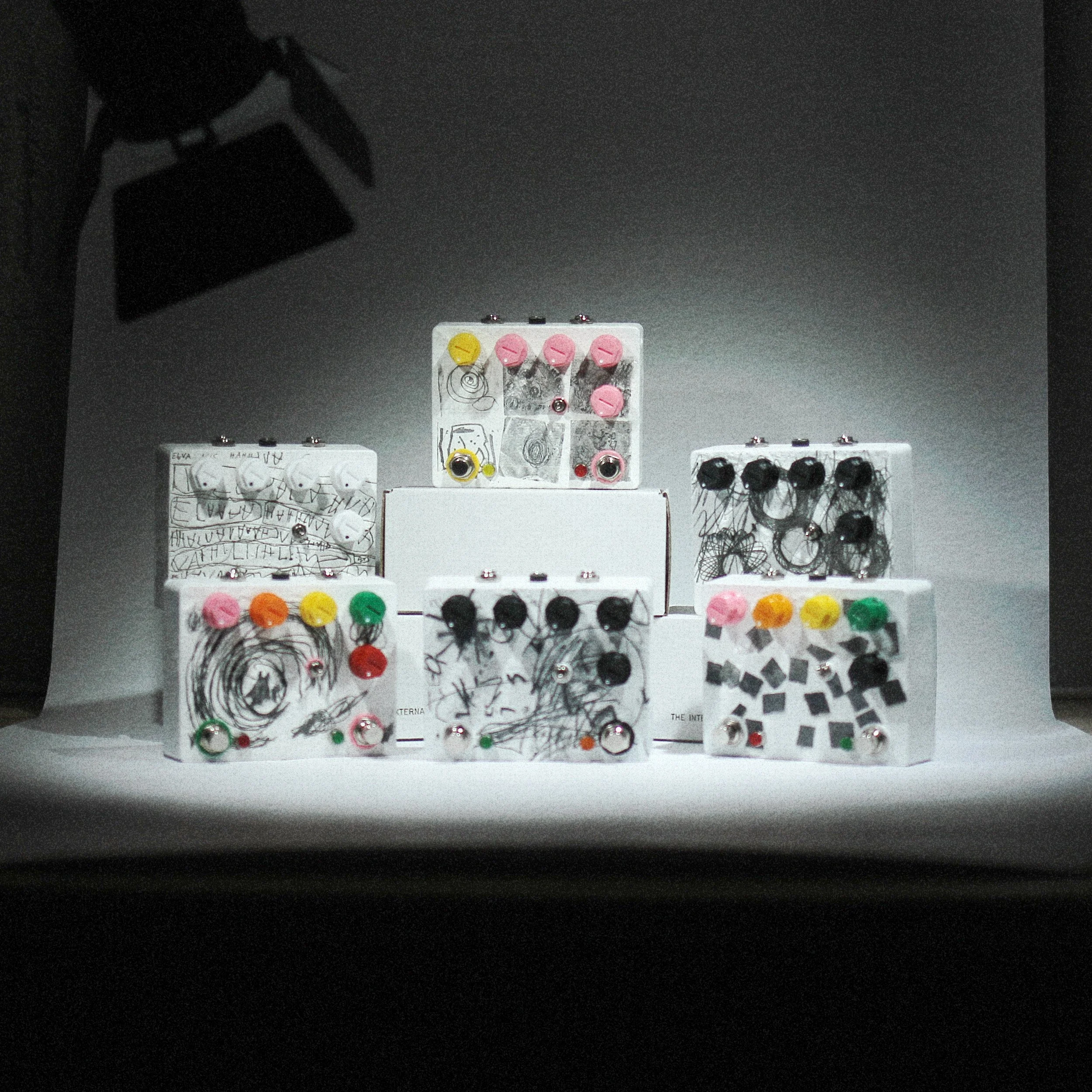 Six white boxes display colorful, handmade electronic circuits with various knobs, wires, and components in an art installation or display setting.