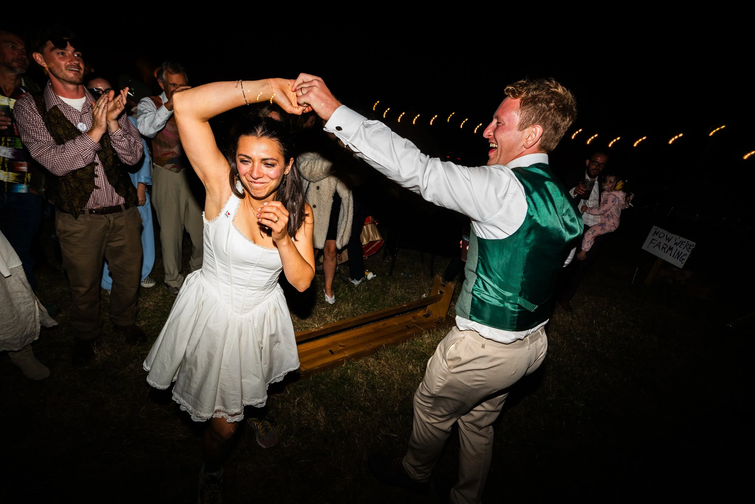 A couple is dancing at a wedding reception, surrounded by guests clapping and smiling, outdoors at night.