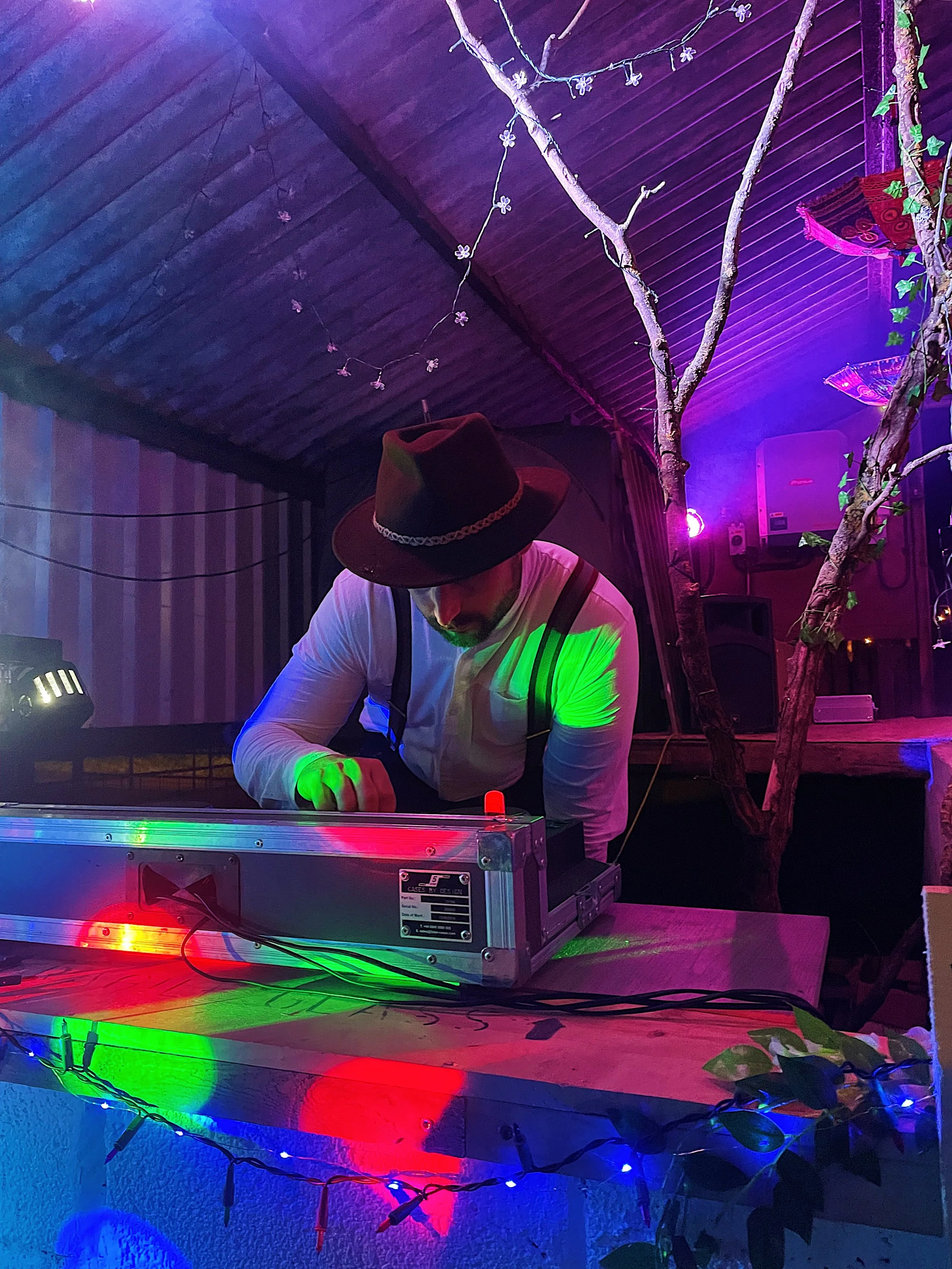 A DJ wearing a hat and suspenders performing at a colorful, decorated venue with tree branches and string lights.