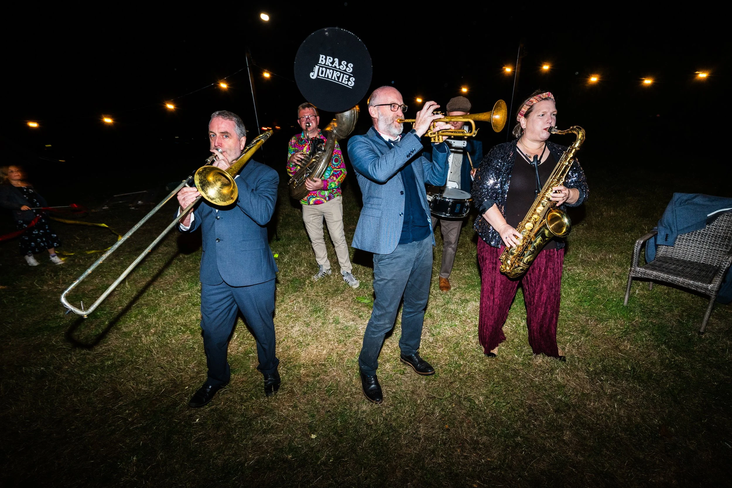 Band of five musicians playing brass and woodwind instruments outdoors at night, under string lights, with a sign reading 'Brass Junkies' in the background.