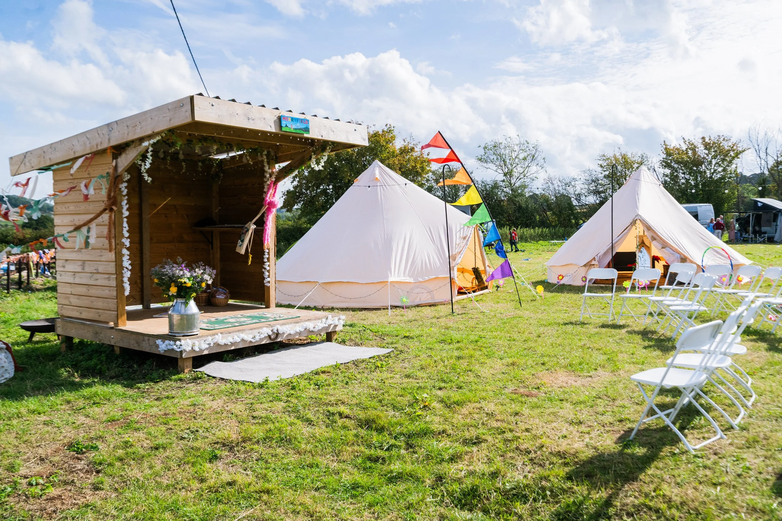 Outdoor camping scene with two white canvas tents, a small wooden stage with flowers in a jug, colorful bunting, folding chairs, and a sunny sky with some clouds.