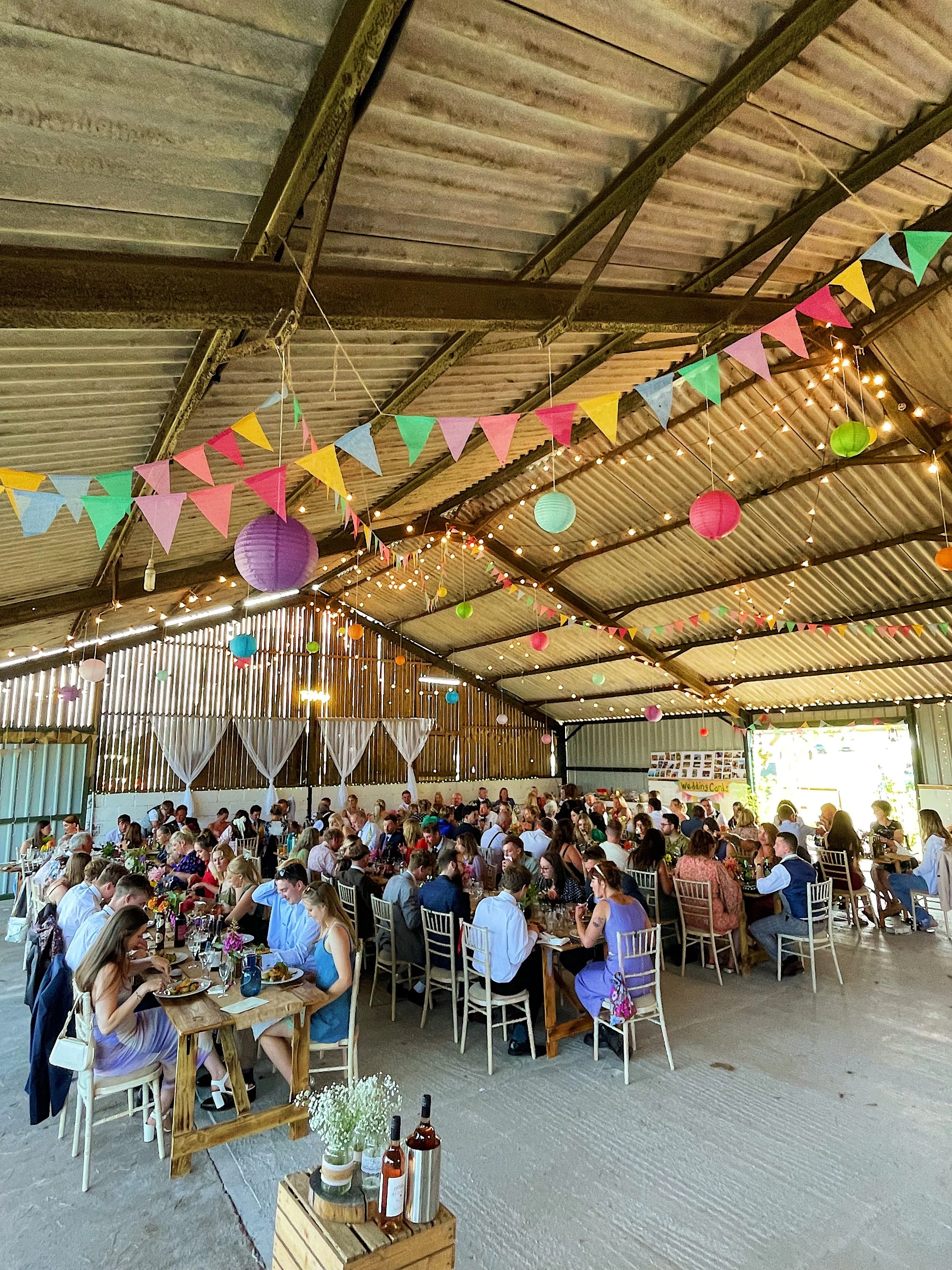 Indoor wedding reception with decorated ceiling featuring colorful bunting and paper lanterns, long tables with guests, and a bright open entrance.