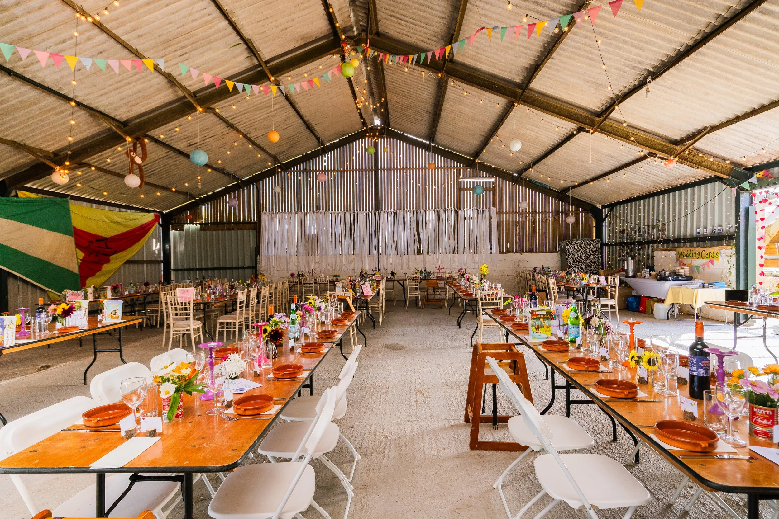 Decorated indoor event space with long tables, colorful flowers, and party decorations such as bunting, lanterns, and fairy lights, set up for a celebration or wedding reception.