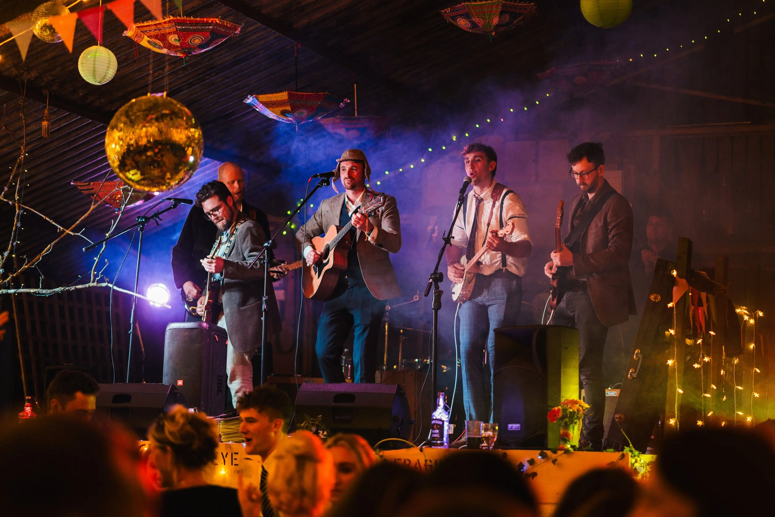 A band of five musicians performing on stage at a lively event with colorful decorations, including paper lanterns and umbrellas, hanging from the ceiling. The stage is decorated with fairy lights and a plant with flowers, and the audience is enjoyin