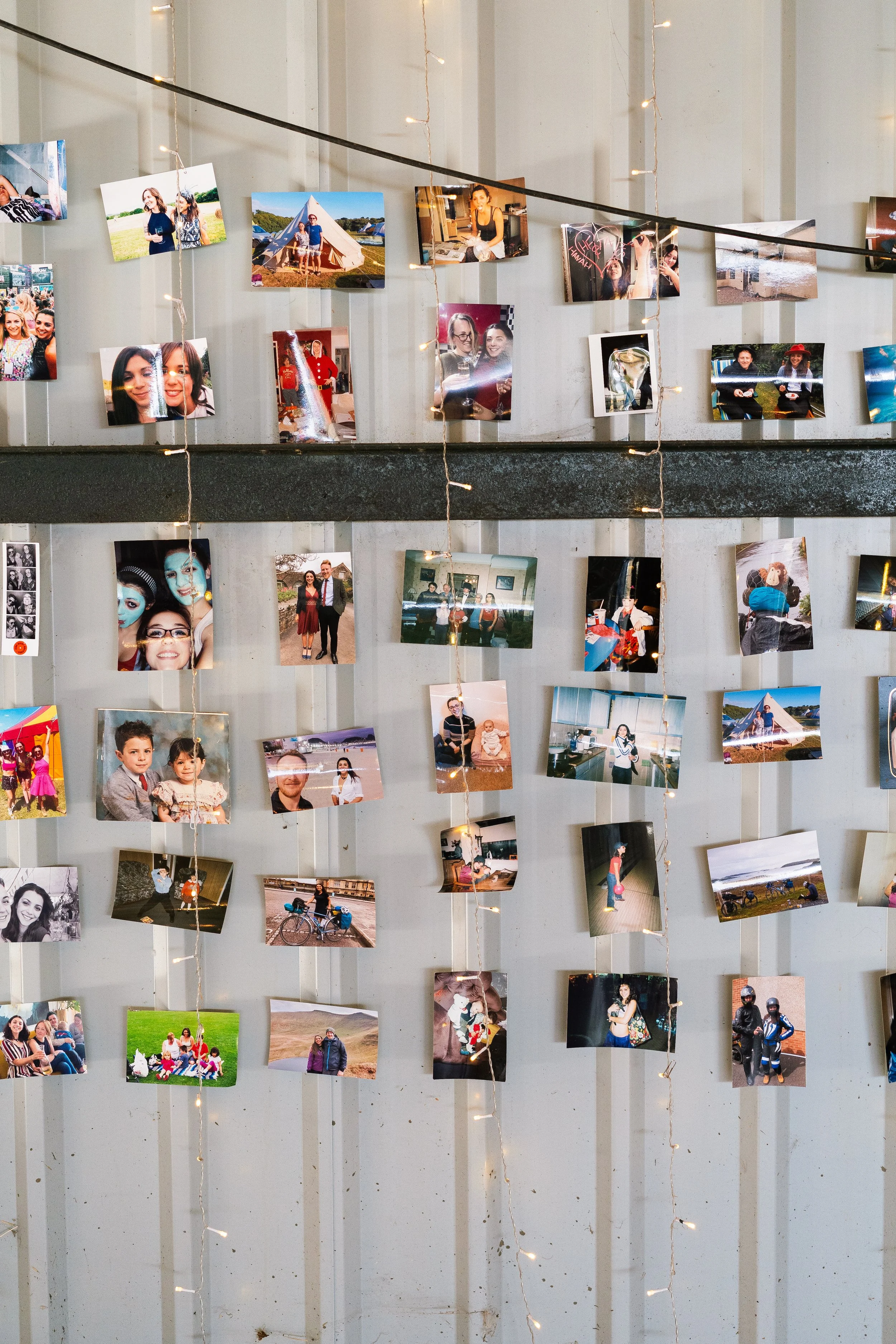 A wall decorated with string lights and numerous printed photos of various people, some with friends or family, some at events, and others outdoors.