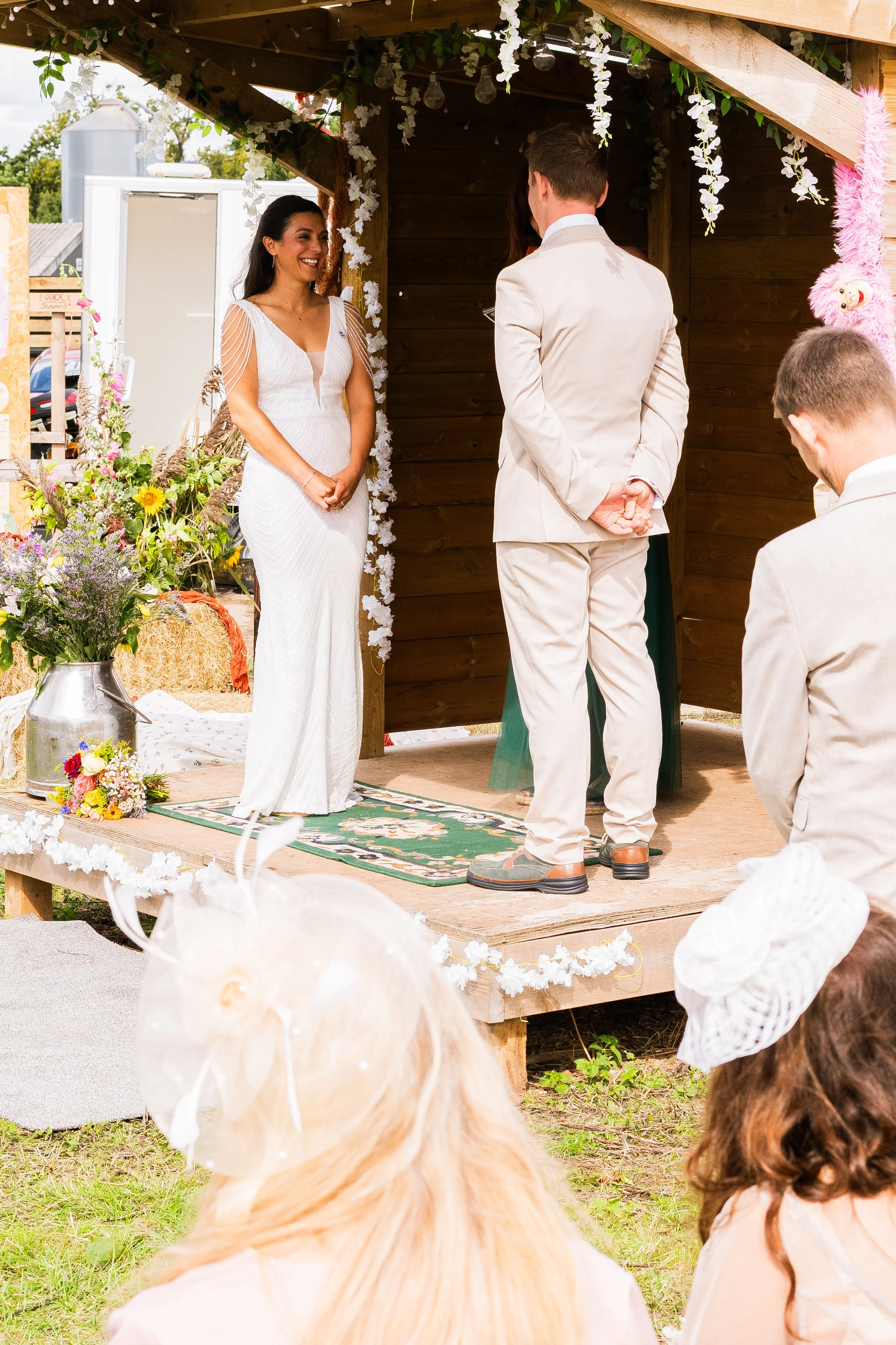 A couple gets married on a small wooden outdoor stage decorated with flowers, with friends and family watching. The bride is wearing a white wedding dress, and the groom is in a light-colored suit. The setting is bright with natural light, decorated 