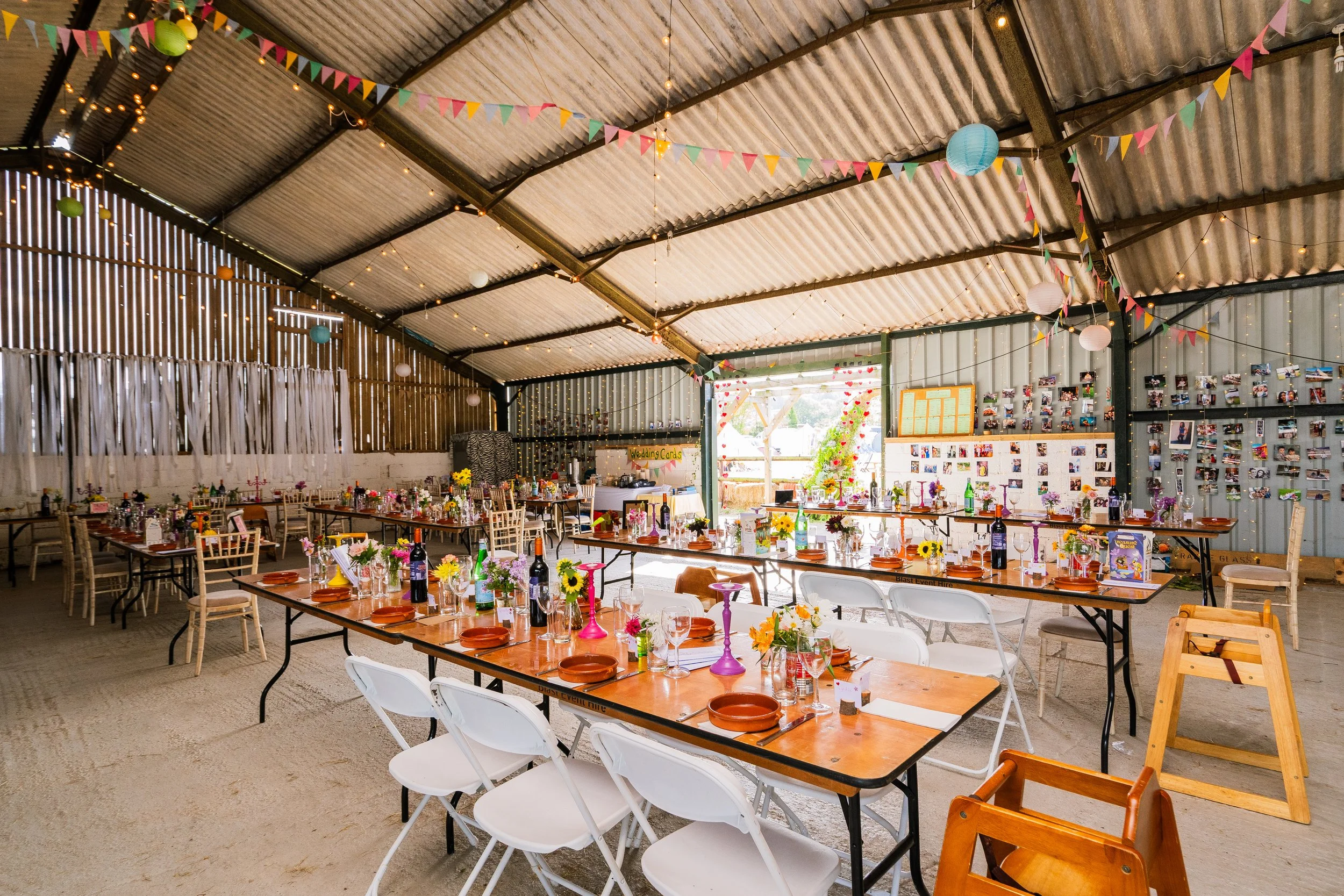 Decorated event space with picnic tables, colorful bunting, paper lanterns, and string lights, set up for a celebration or wedding reception.