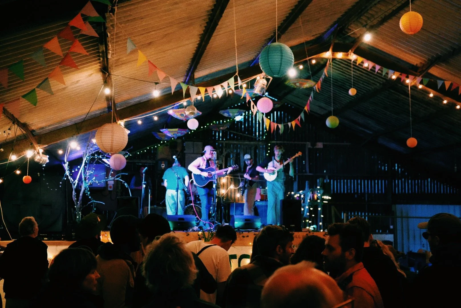 Live band performing on stage decorated with colorful bunting, paper lanterns, and string lights, with an audience watching inside a rustic venue.
